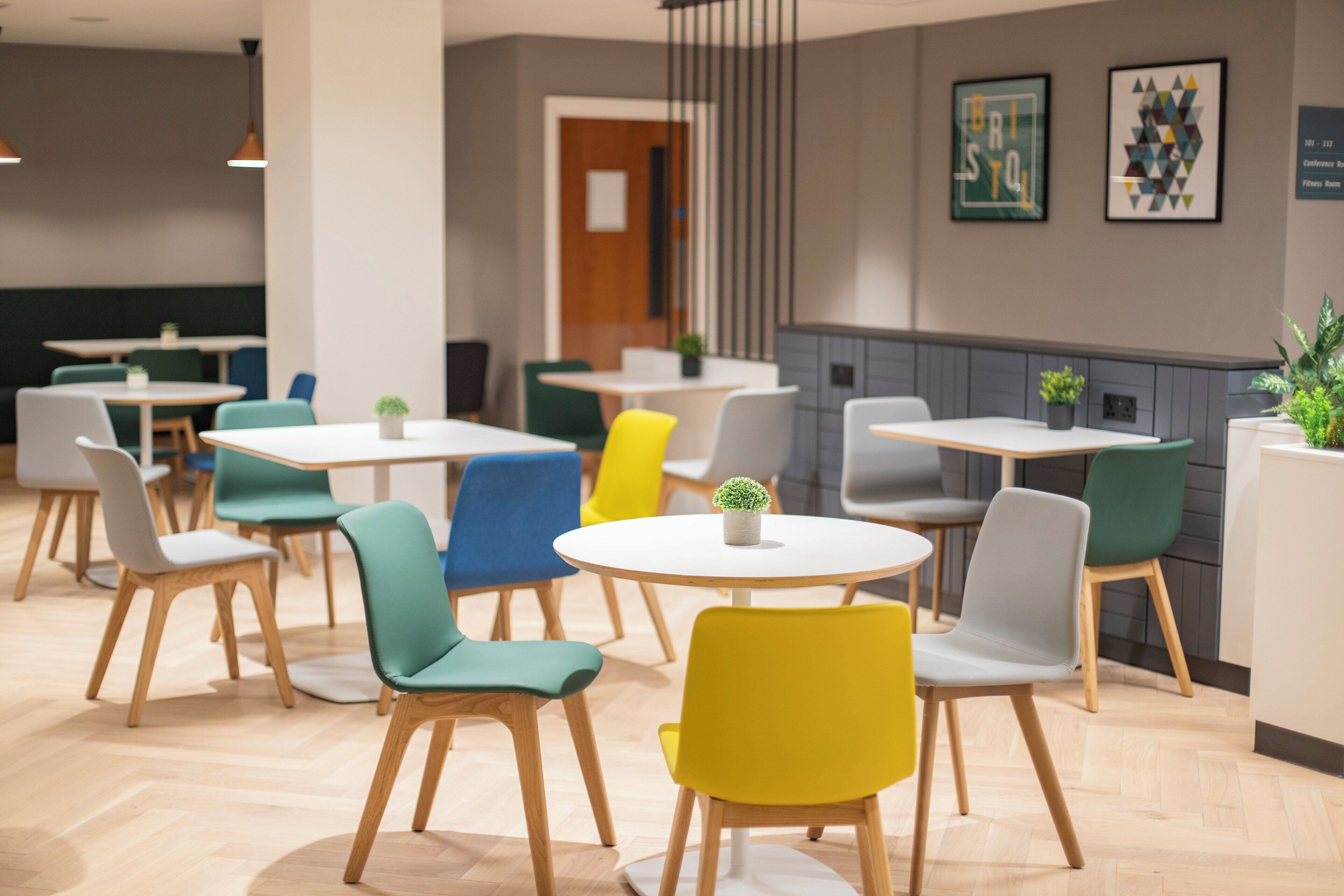 Un comedor moderno con mesas y sillas redondas en varios colores, suelo de madera y una pared con obras de arte enmarcadas.