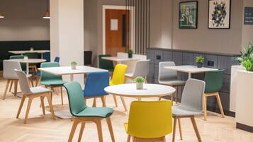 Un comedor moderno con mesas y sillas redondas en varios colores, suelo de madera y una pared con obras de arte enmarcadas.