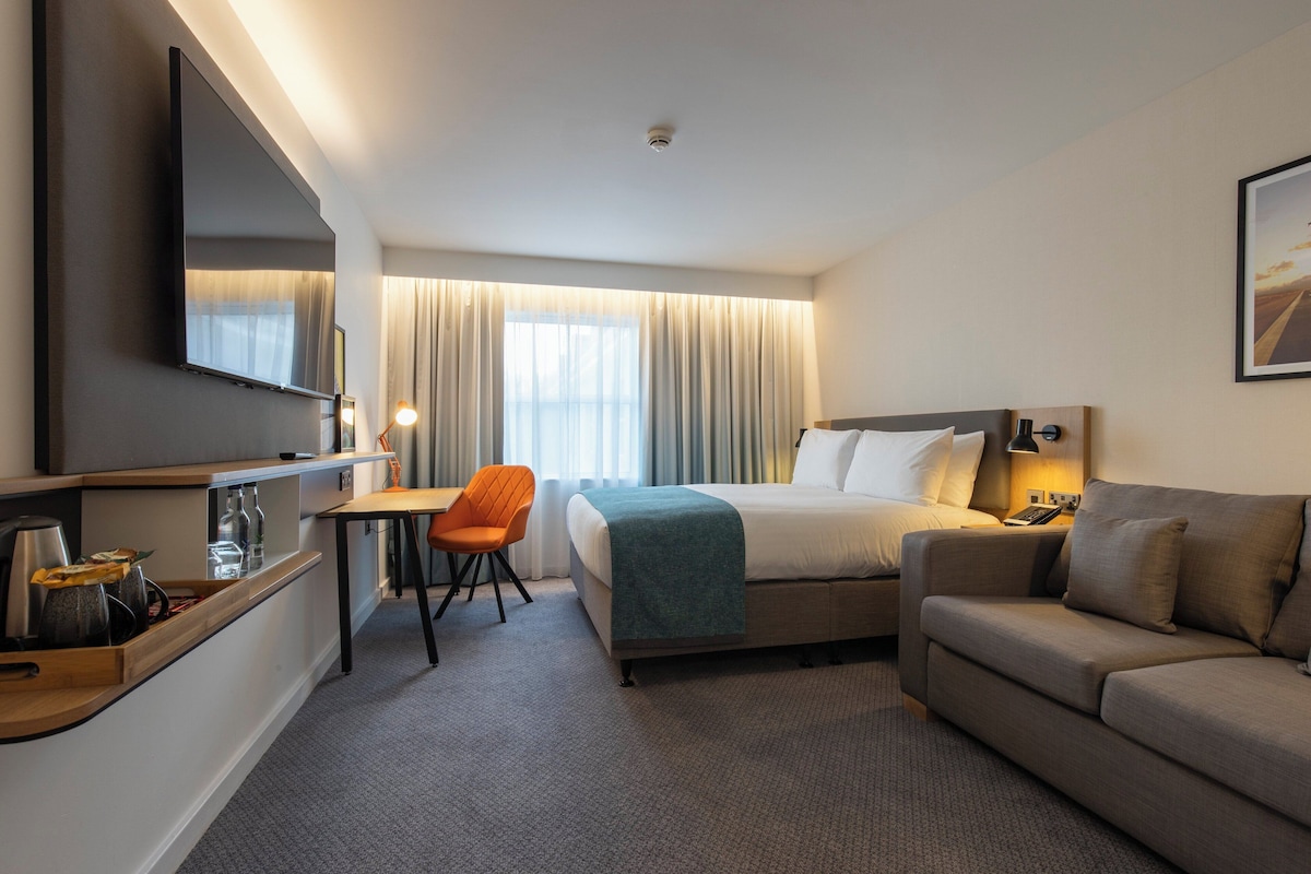 Een hotelkamer met een bed, een slaapbank, een bureau met stoel, een aan de muur bevestigde televisie en een raam met gordijnen.