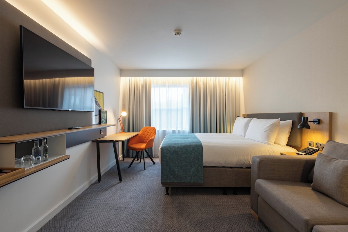 A hotel room with a bed, a desk, a chair, a TV, and a bedside table.