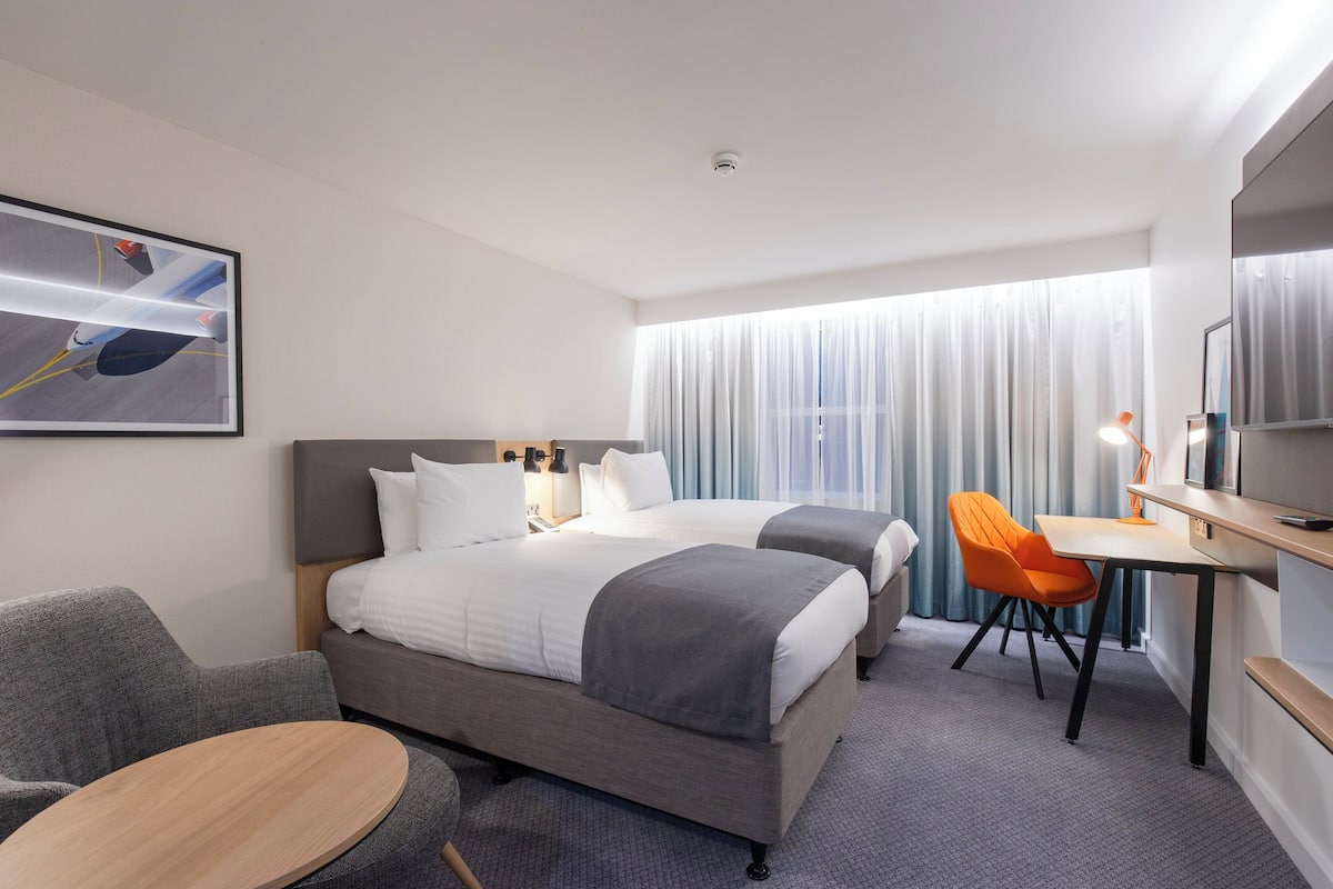 A hotel room with two beds, a desk, a chair, a TV, and a painting on the wall.