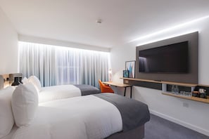 Habitación de hotel con dos camas, televisión, escritorio con silla y estante con objetos.
