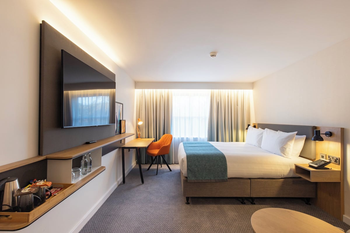 A hotel room with a bed, a desk, a chair, a TV, and a bedside table with a lamp.