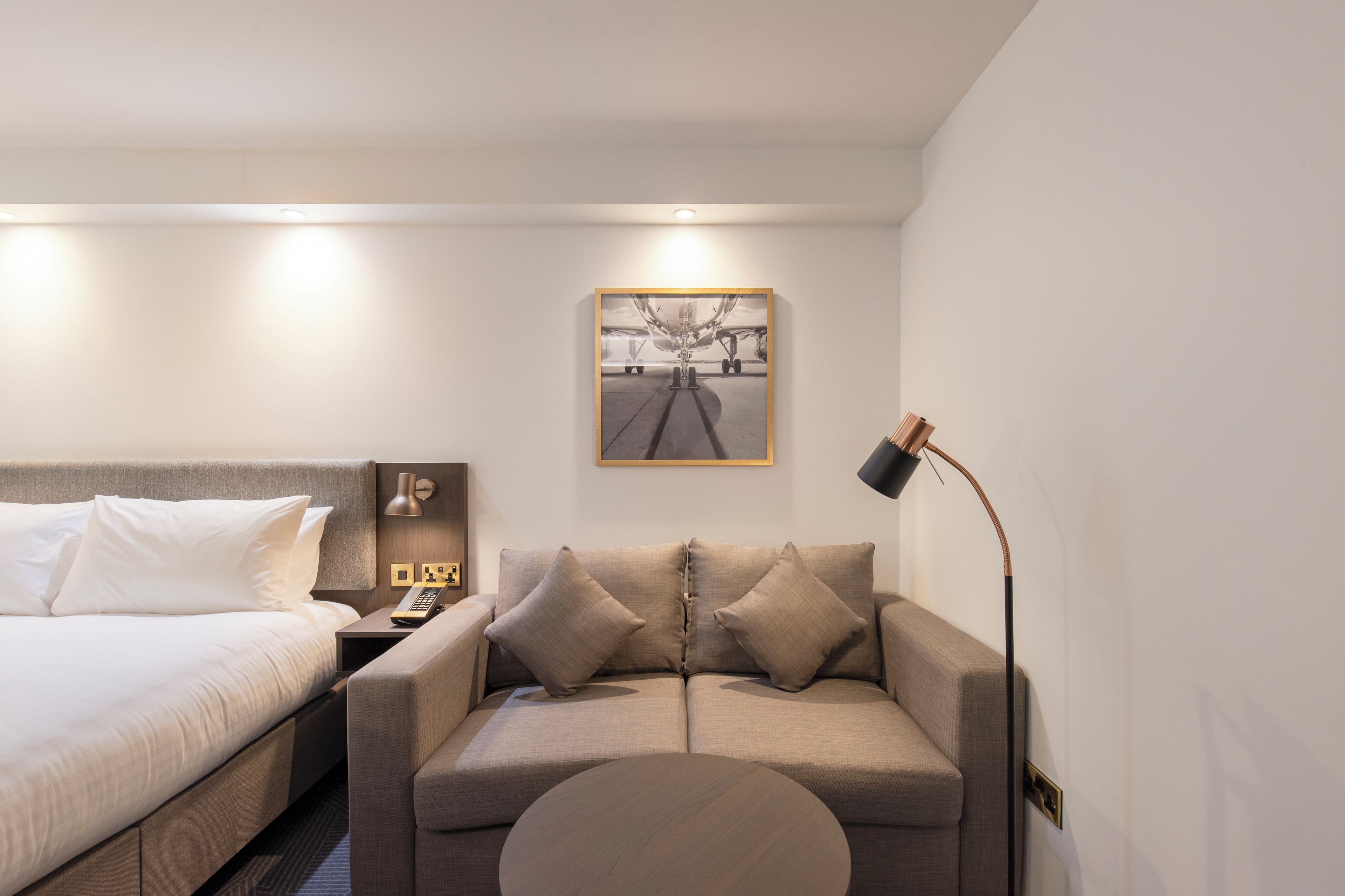 A modern hotel room with a bed, a sofa, a desk with a lamp, and a painting on the wall.
