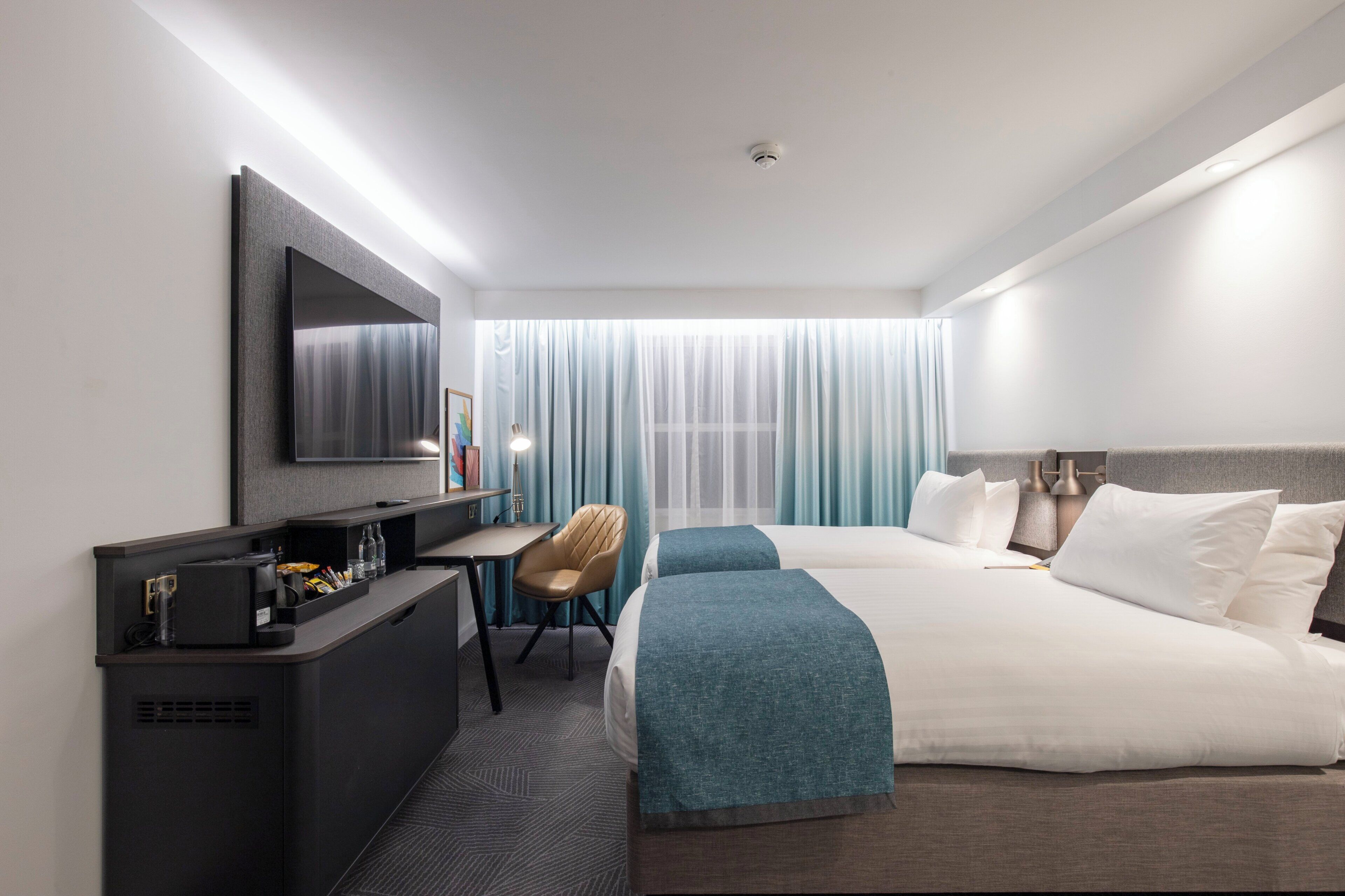 A hotel room with a bed, a desk with a chair, a TV, and a window with curtains.