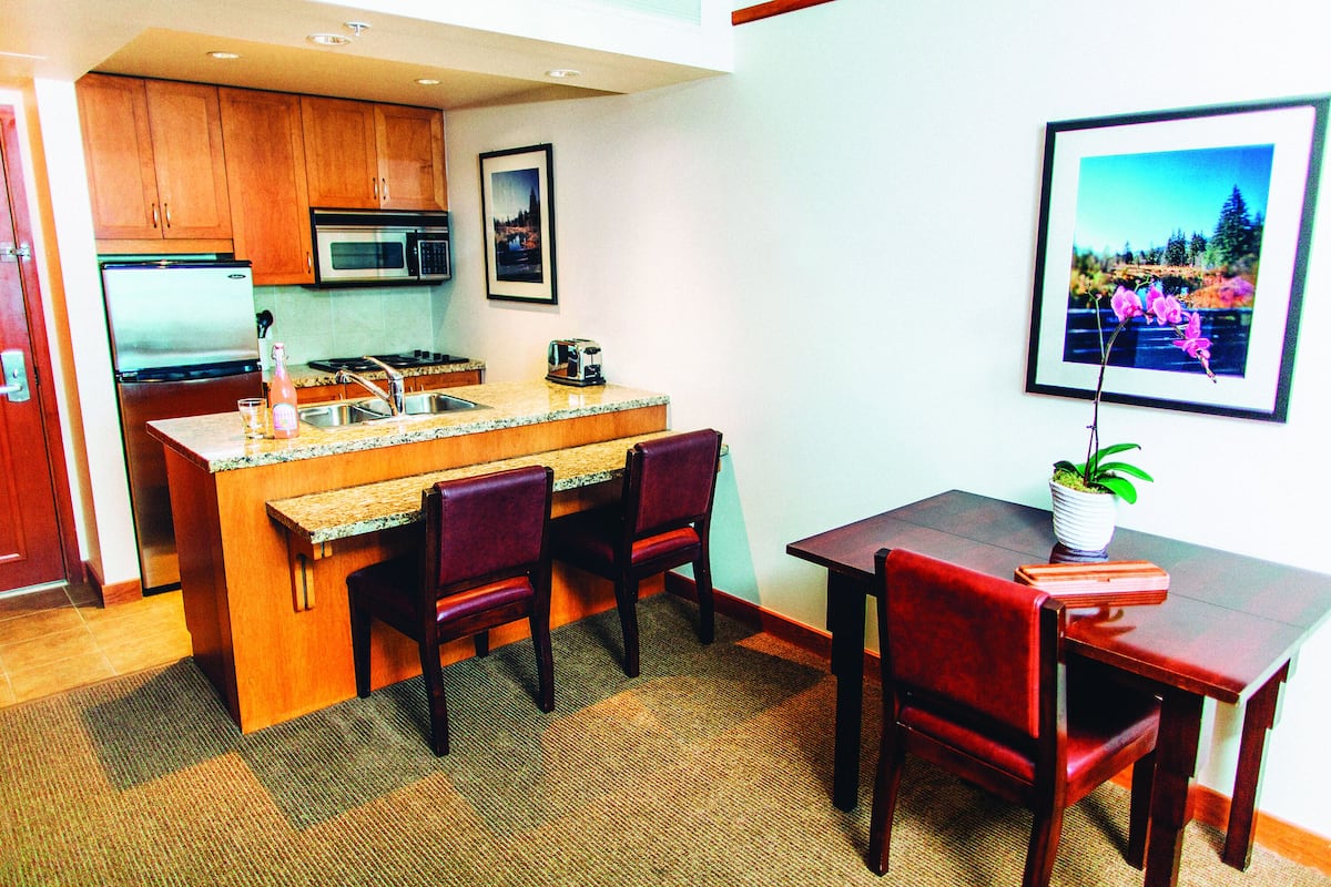 A compact kitchen with wooden cabinets, a microwave, and a sink. There is a dining area with a table and chairs, and a framed picture on the wall.