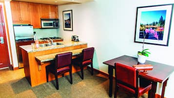 A compact kitchen with wooden cabinets, a microwave, and a sink. There is a dining area with a table and chairs, and a framed picture on the wall.