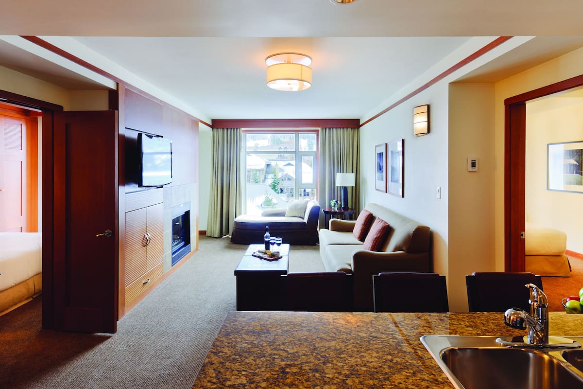 A hotel room with a living area featuring a sofa, a coffee table, a TV, and a dining area with a sink and a granite countertop.