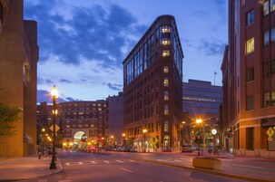 A city street at dusk with modern buildings, street lamps, and a pedestrian crossing.