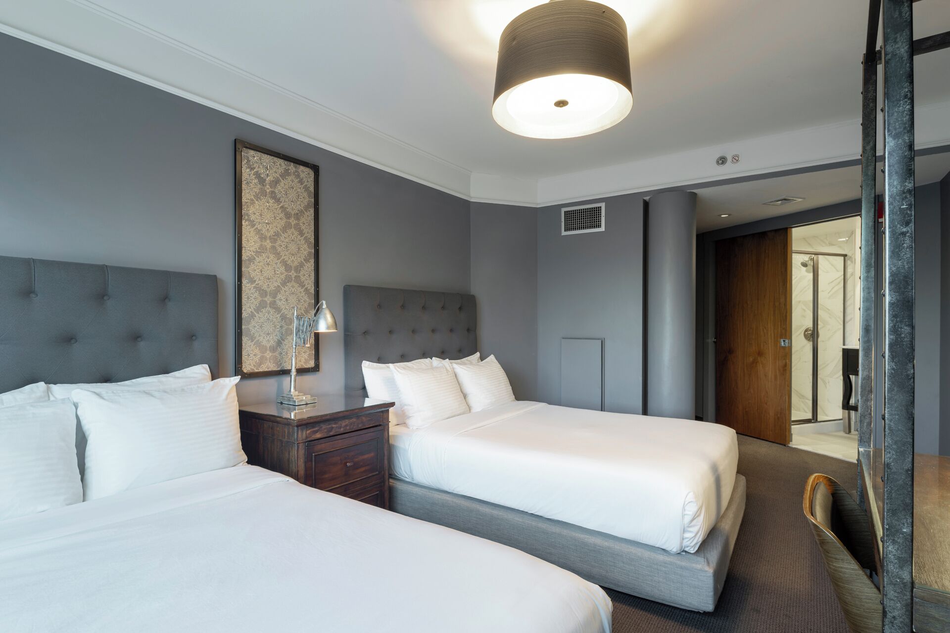 A hotel room with two beds, a nightstand, a lamp, a large mirror, and a door leading to a bathroom.