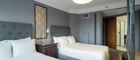 A hotel room with two beds, a nightstand, a lamp, a large mirror, and a door leading to a bathroom.