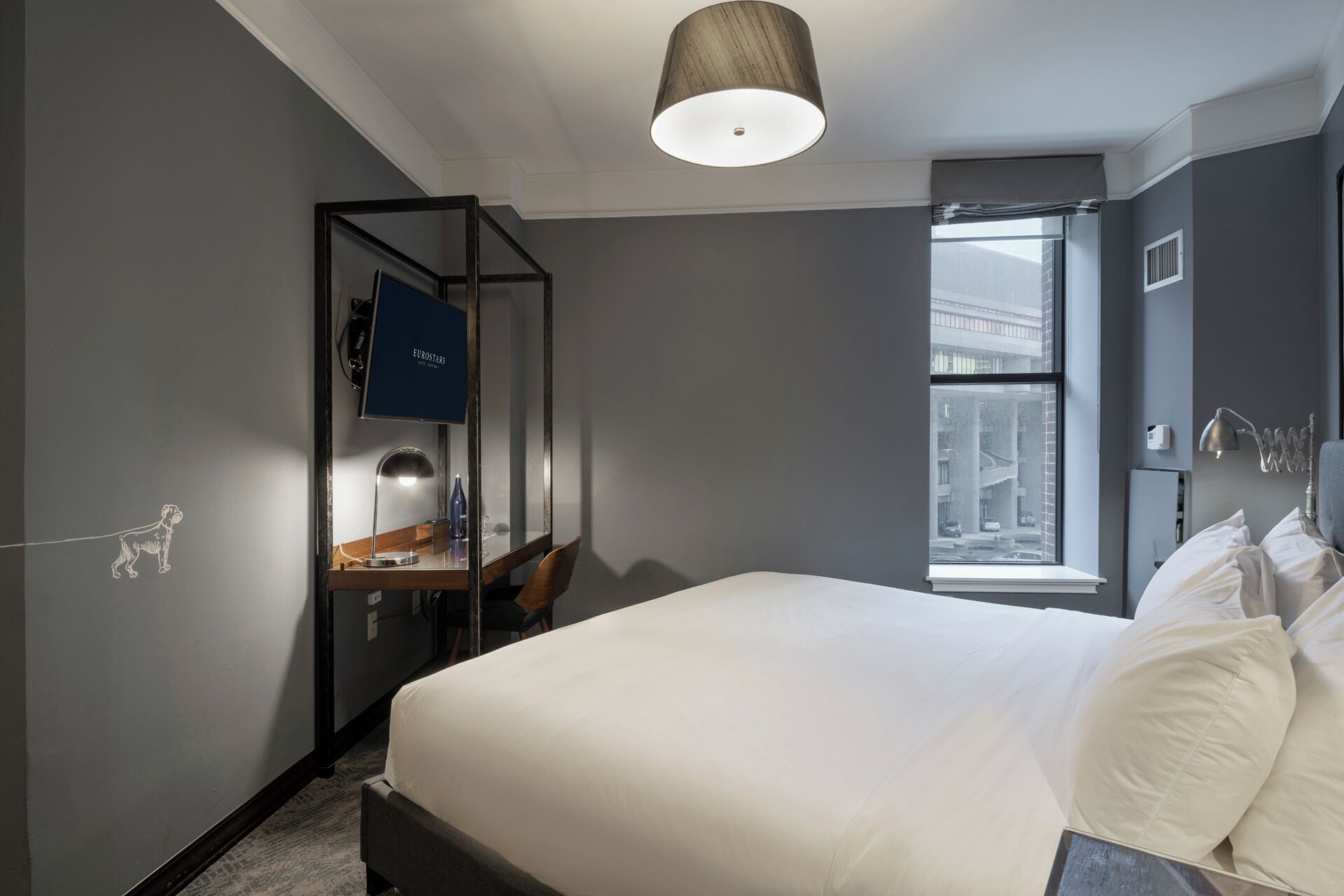 A modern hotel room with a large bed, a desk with a lamp, a window with a view of buildings, and a ceiling light.