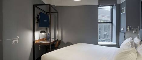 A modern hotel room with a large bed, a desk with a lamp, a window with a view of buildings, and a ceiling light.