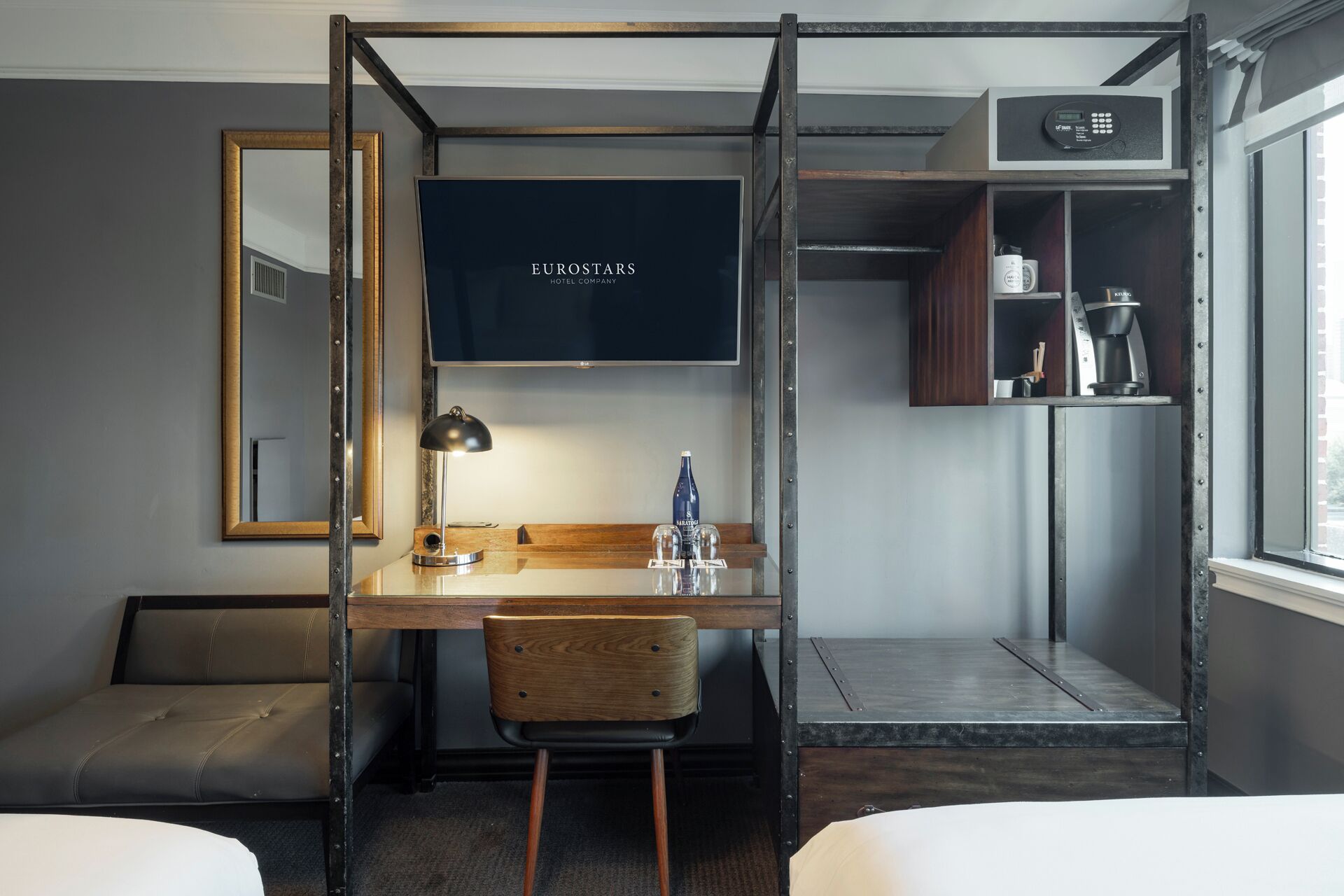 A compact hotel room with a bed, a desk with a chair, a mirror, a TV screen displaying 'EUROSTARS HOTEL COMPANY', a shelf with toiletries, and a window with blinds.