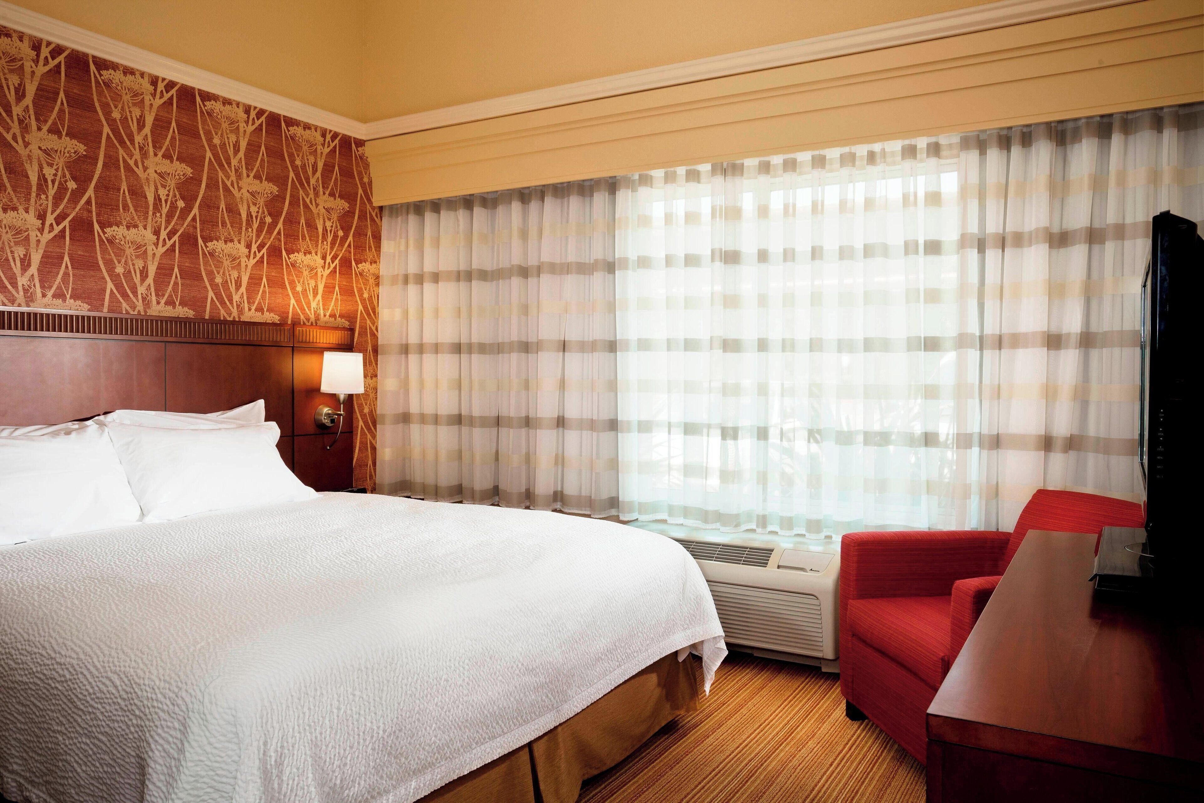 A hotel room with a large bed, a red armchair, a television, and a window with striped curtains.