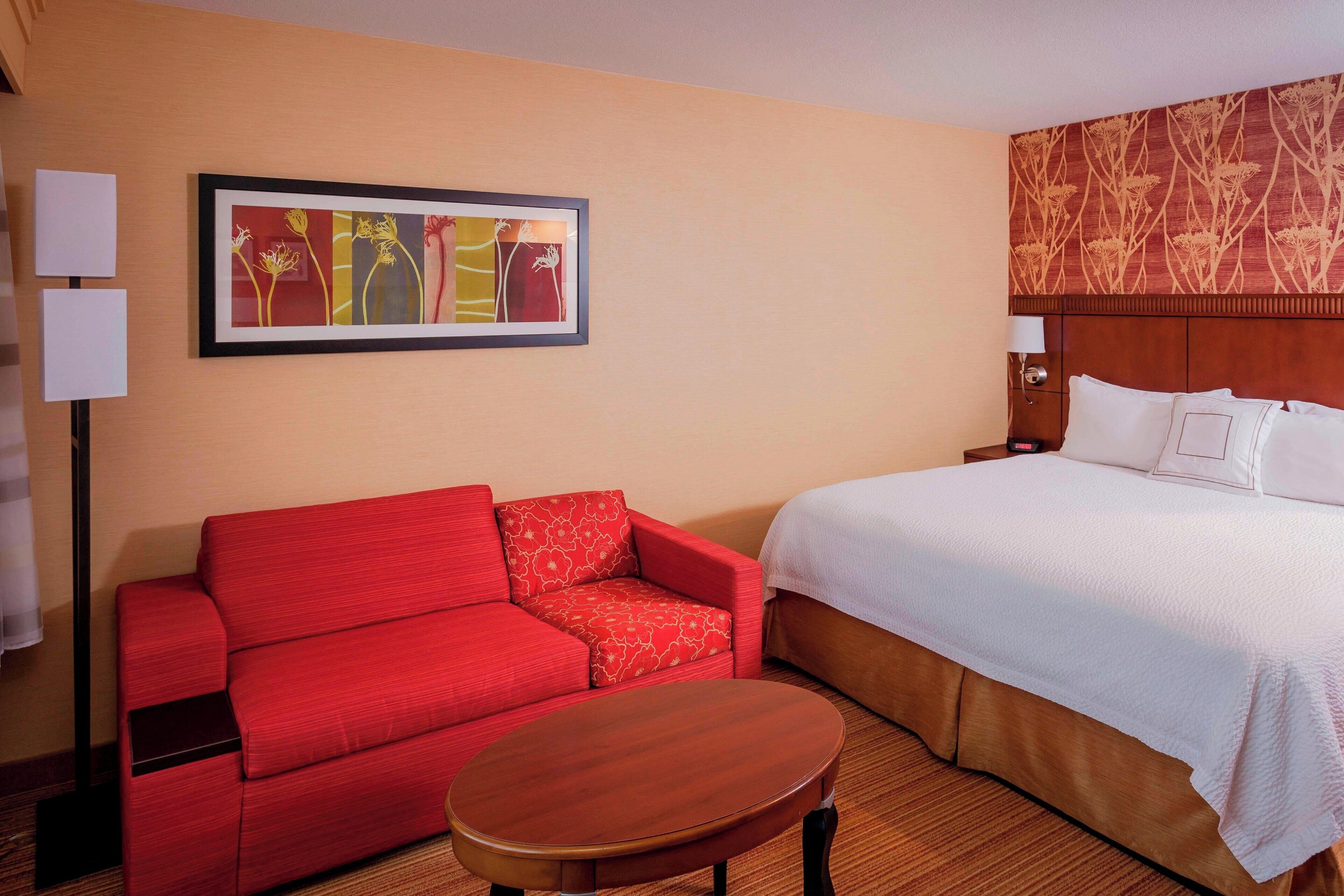 A hotel room with a red sofa, a wooden table, a large bed with white bedding, and a framed abstract painting on the wall.