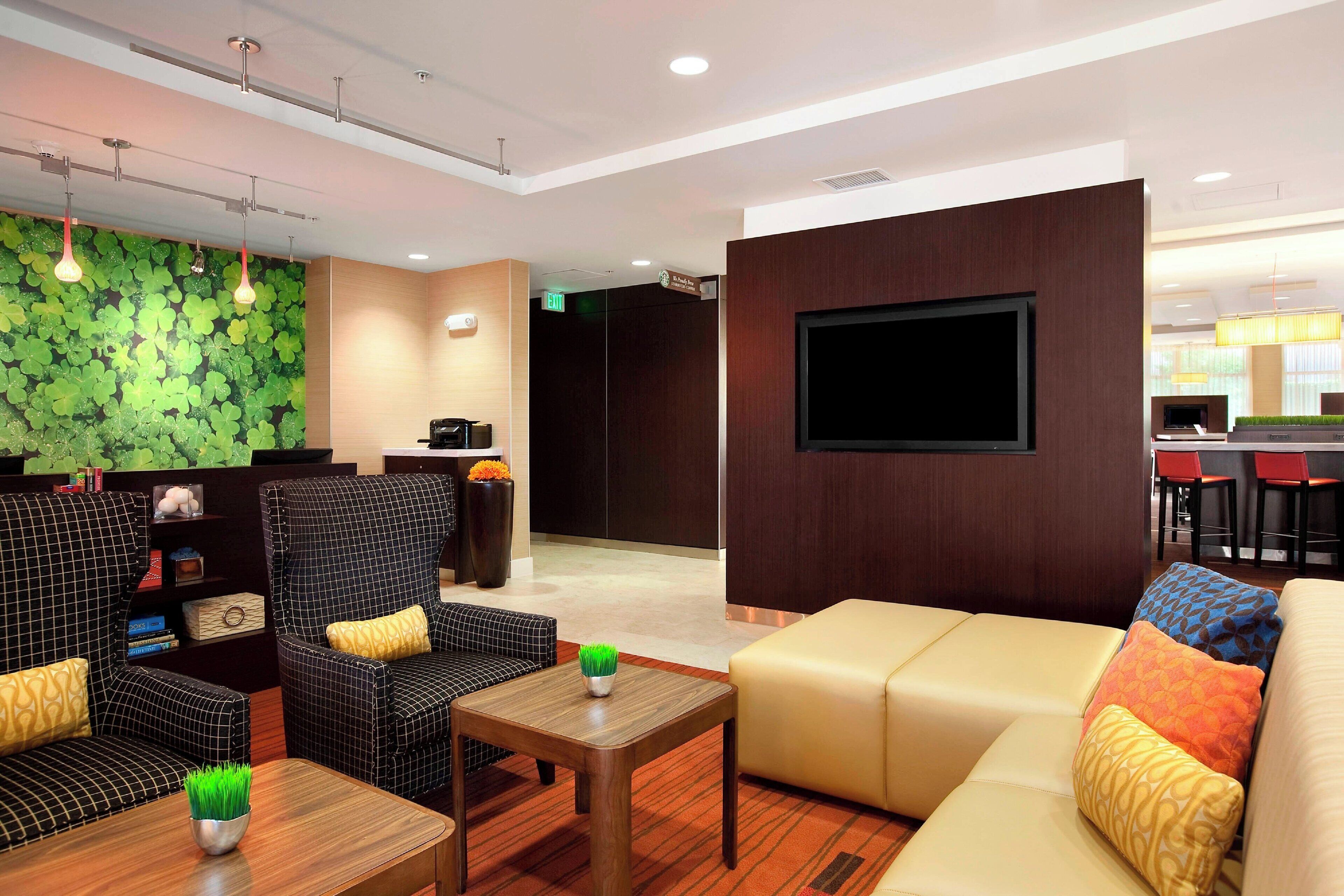 A modern lobby with a green leaf wall, seating areas, a reception desk, and a TV screen.