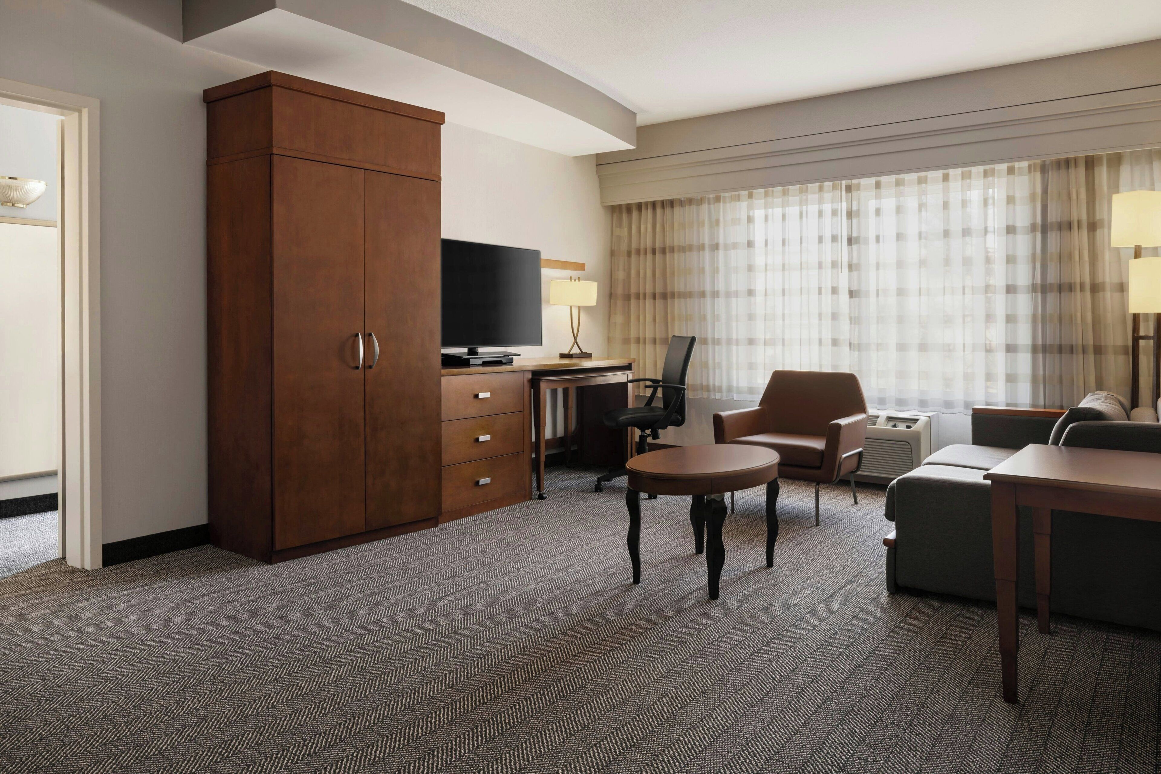 A hotel room with a large wooden wardrobe, a flat-screen TV, a desk with a lamp, a sofa, and a chair.