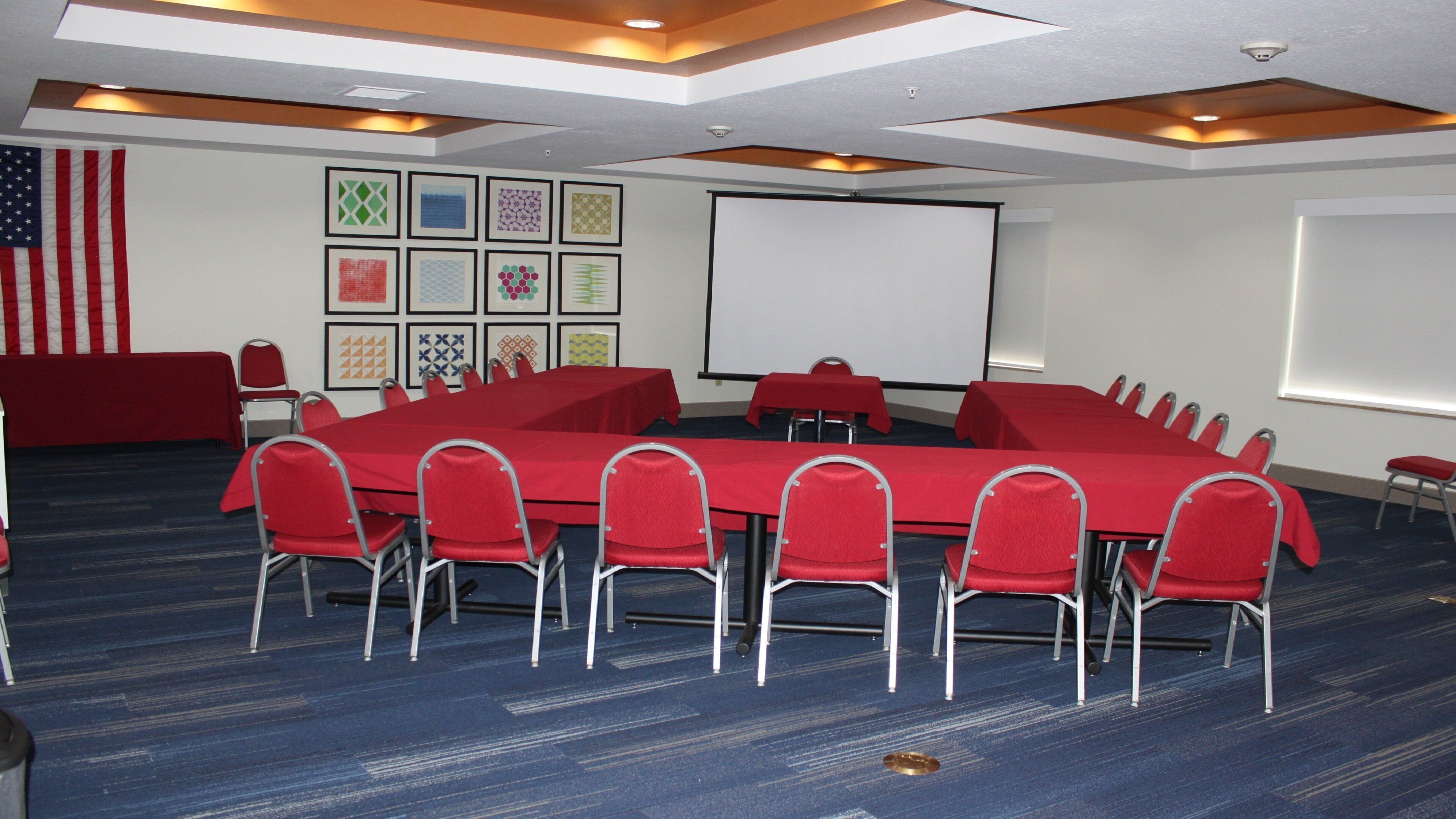A conference room with a long table, red chairs, a projector screen, and a wall with patterned artwork.