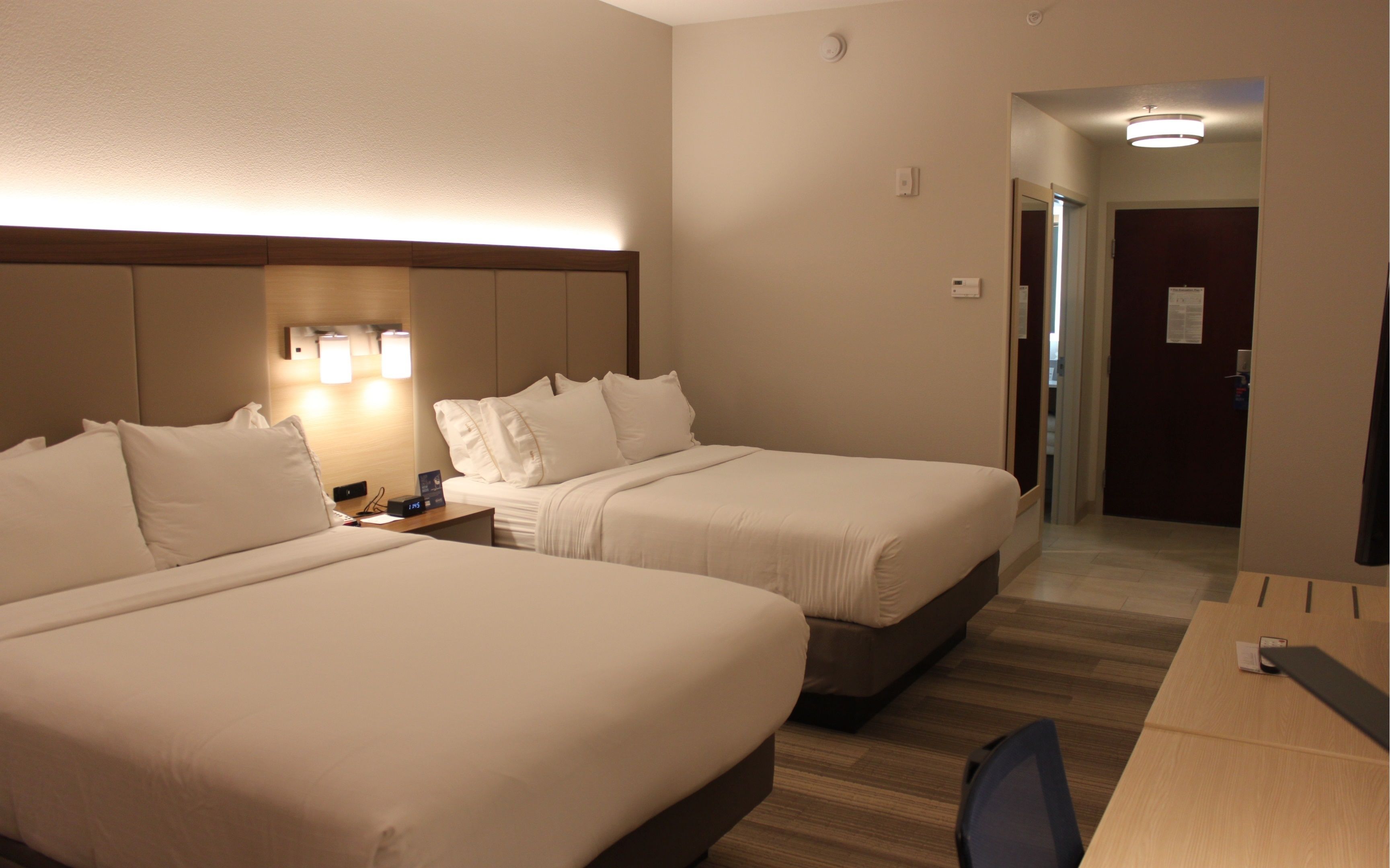 A hotel room with two beds, a desk, and a chair. There is a nightstand with a clock and a lamp.