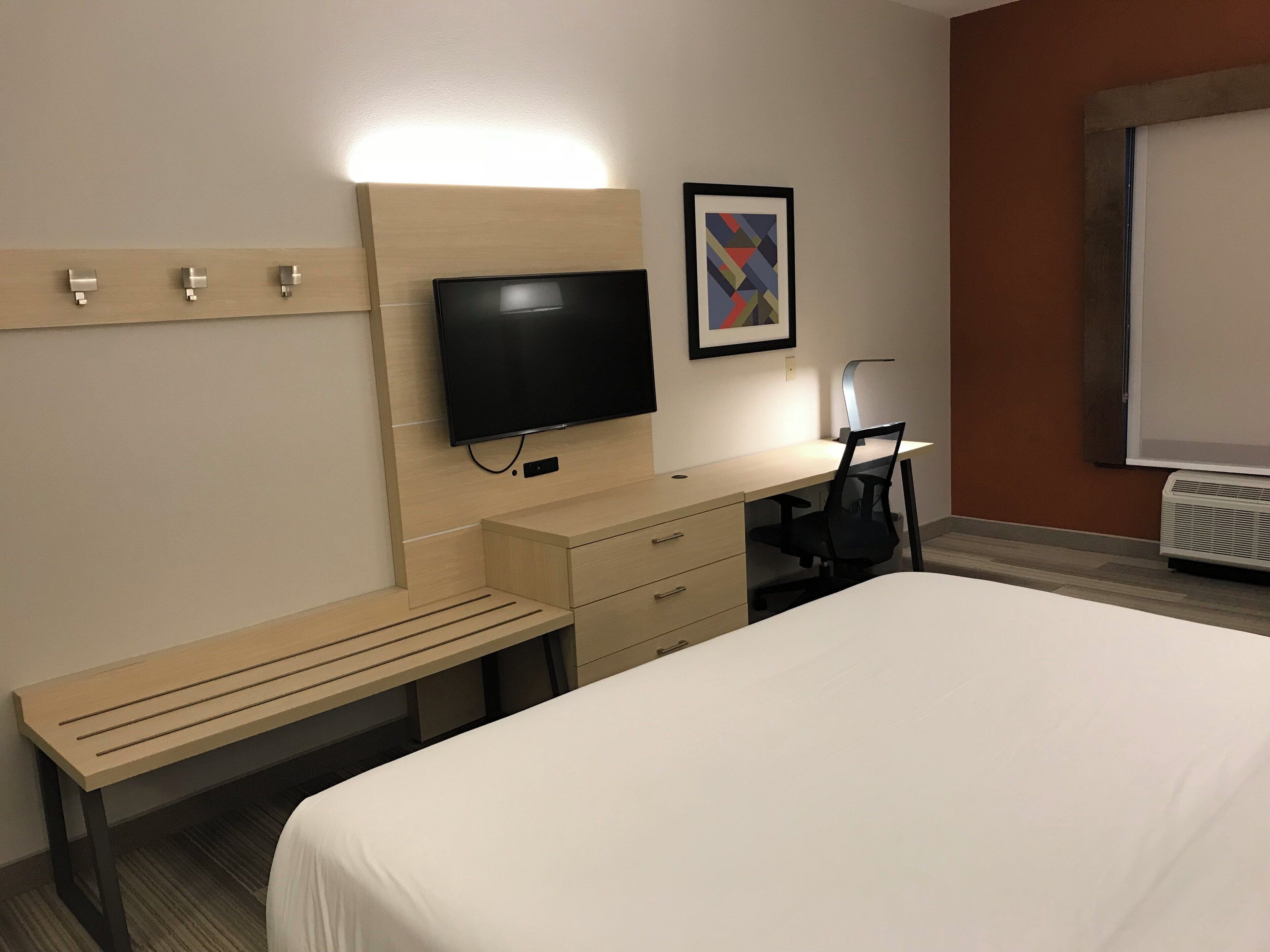 A hotel room with a bed, a desk, a chair, a television, a bench, and a framed picture on the wall.