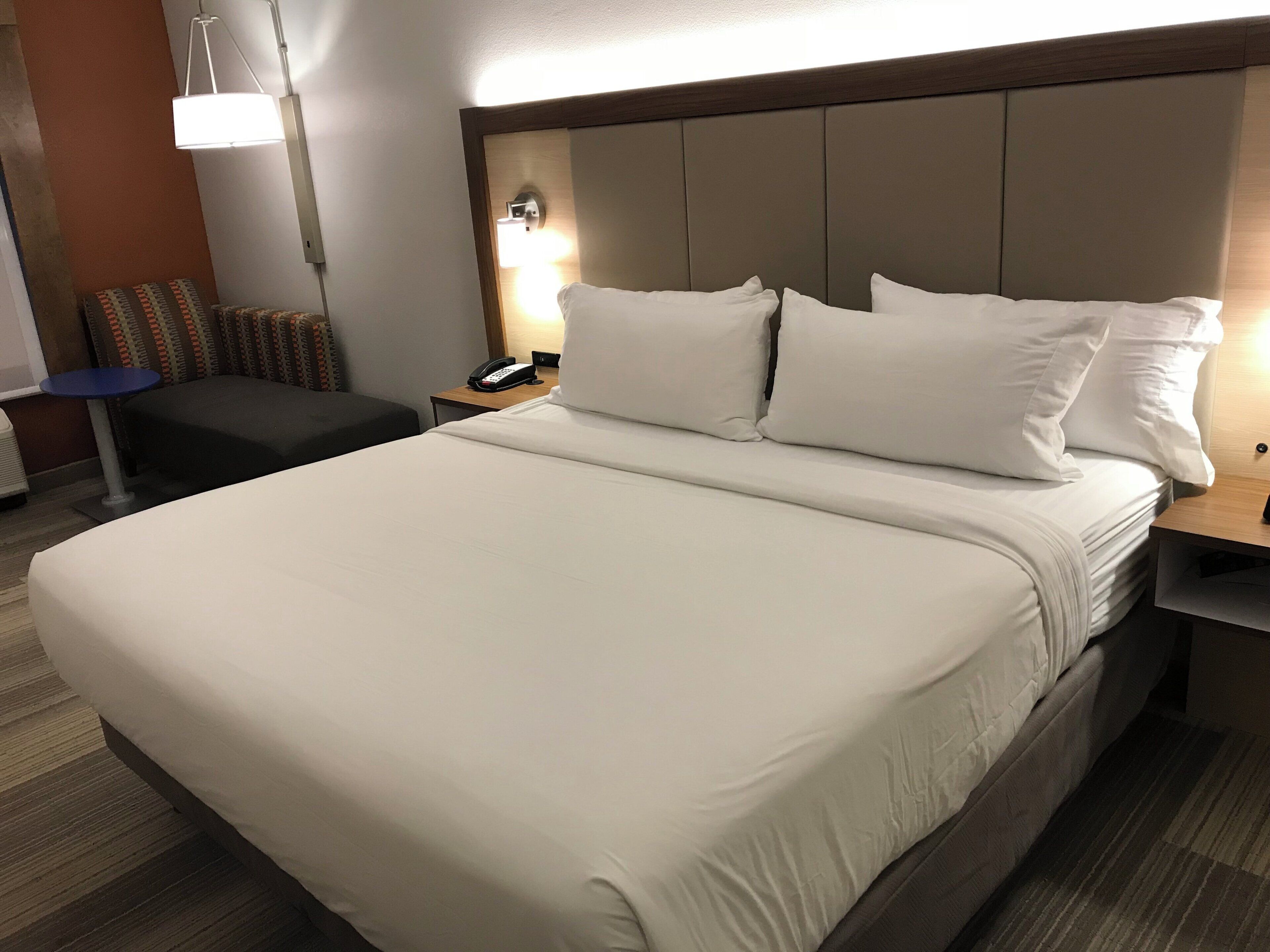 A hotel room with a large bed, a bedside table with a lamp, a small table with a phone, and a chair.