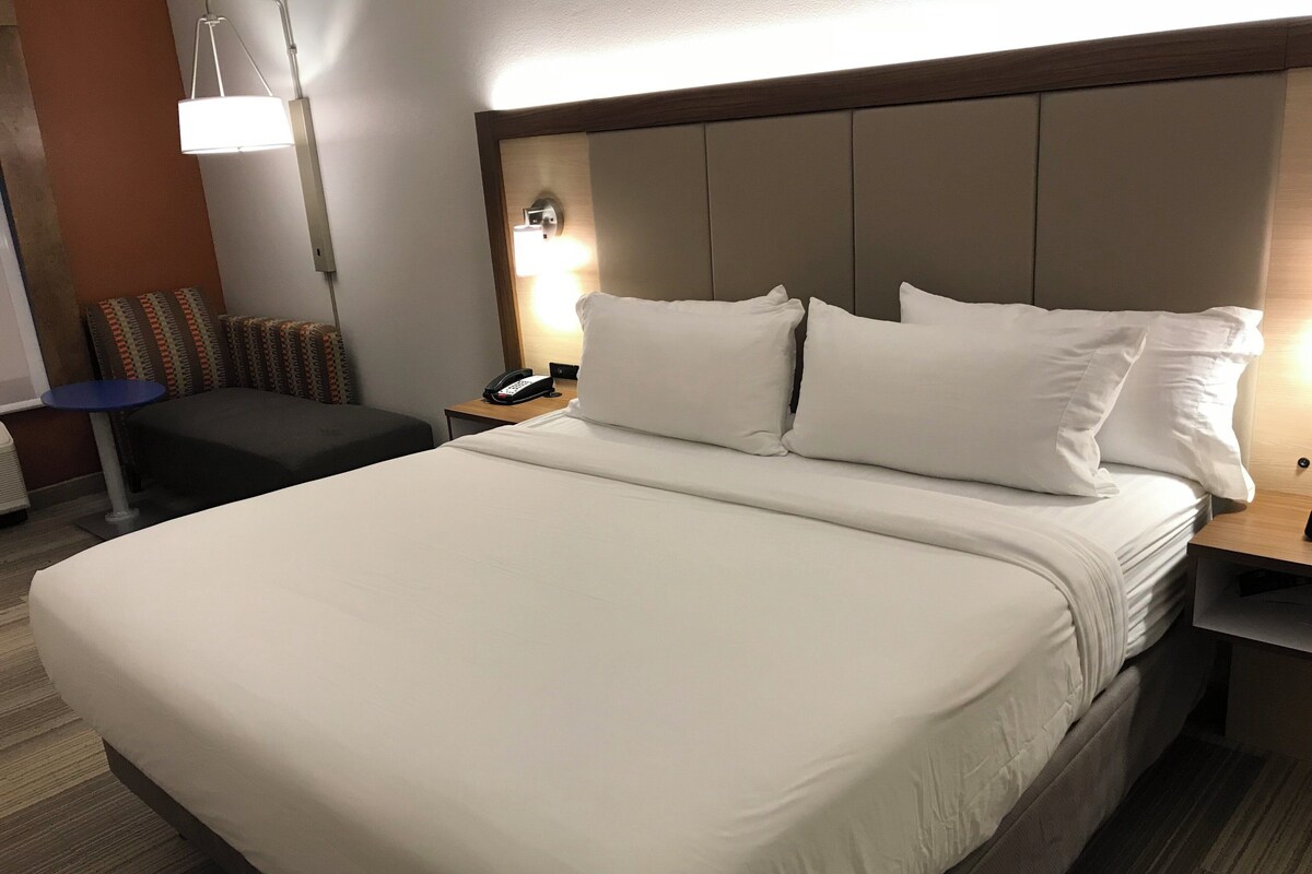A hotel room with a large bed, a bedside table with a lamp, a small table with a phone, and a chair.