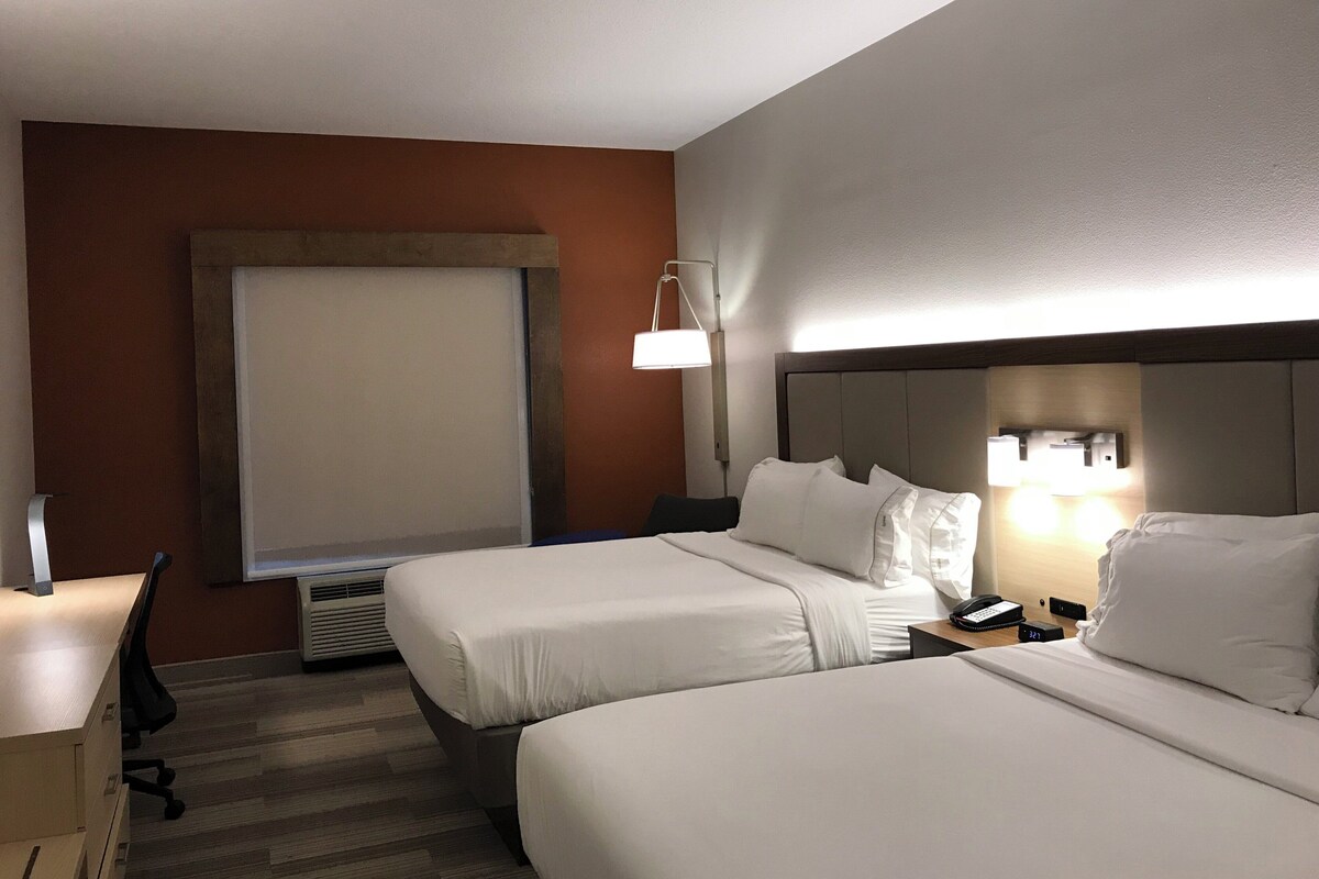 A hotel room with two beds, a desk, a chair, a lamp, and a window.