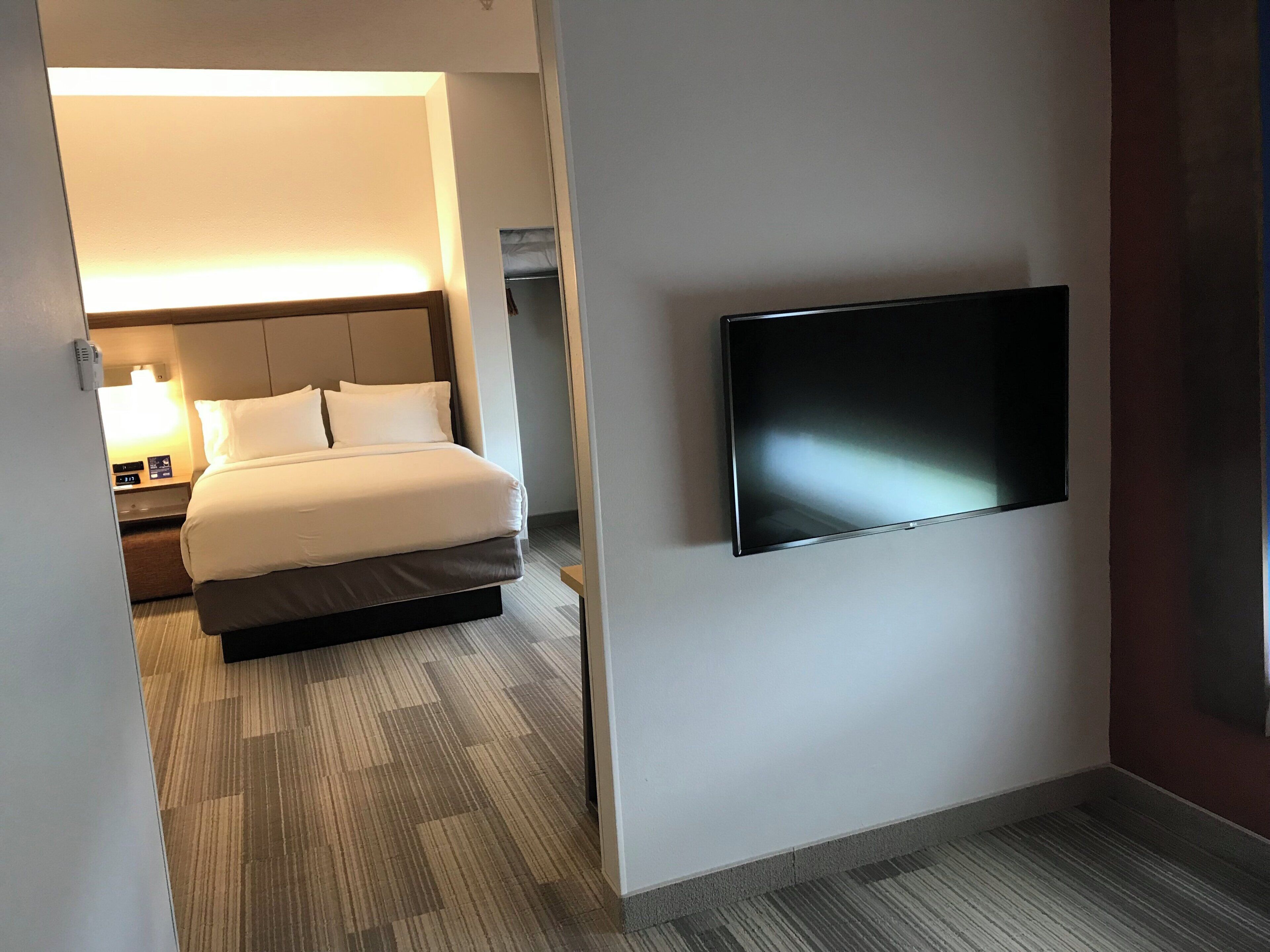 Ett hotellrum med en säng, ett sängbord med en lampa, en platt-TV som är monterad på väggen och ett golv med heltäckningsmatta.