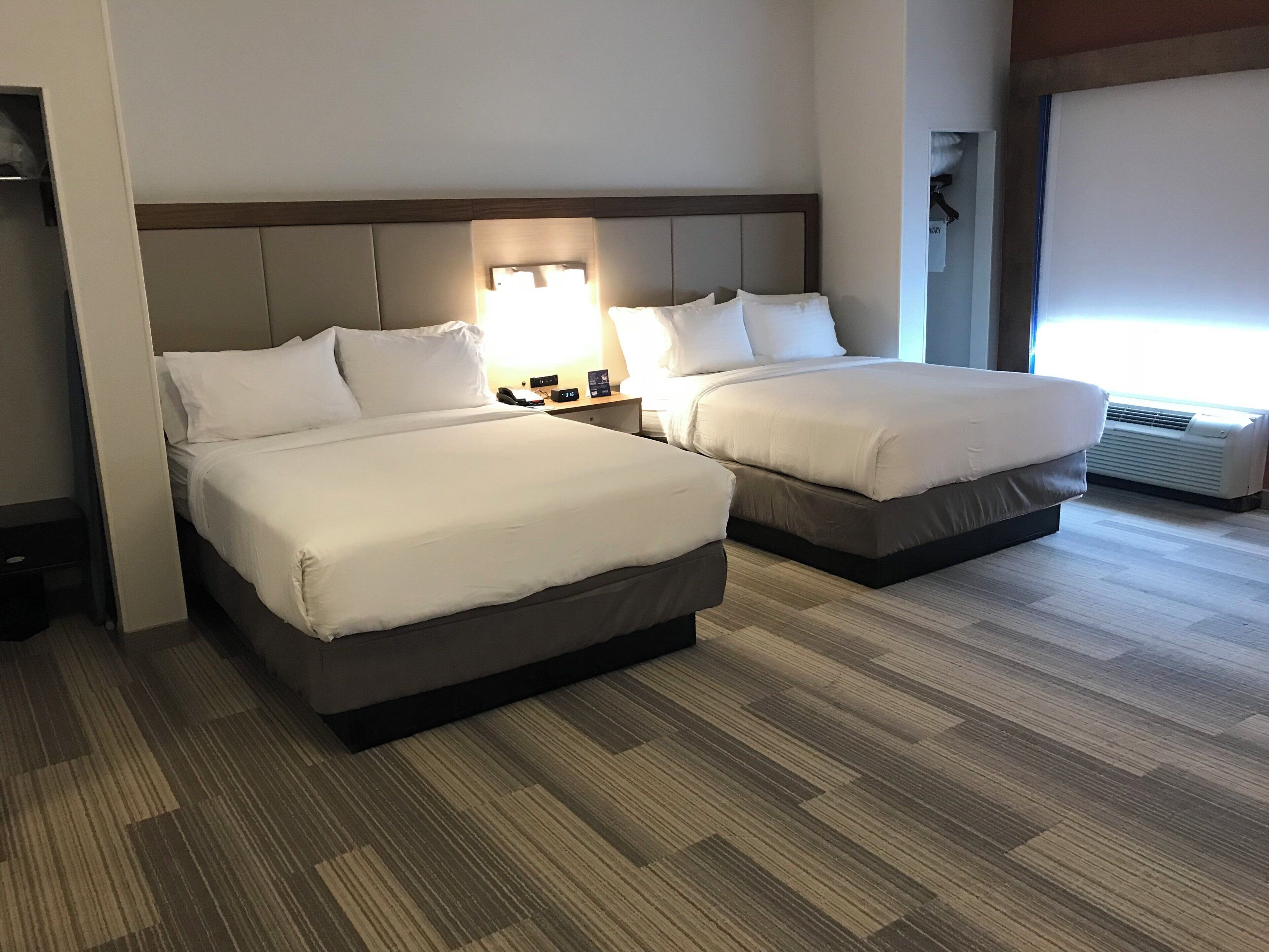A hotel room with two beds, a nightstand with a lamp, a wall-mounted air conditioner, and a window with blinds.