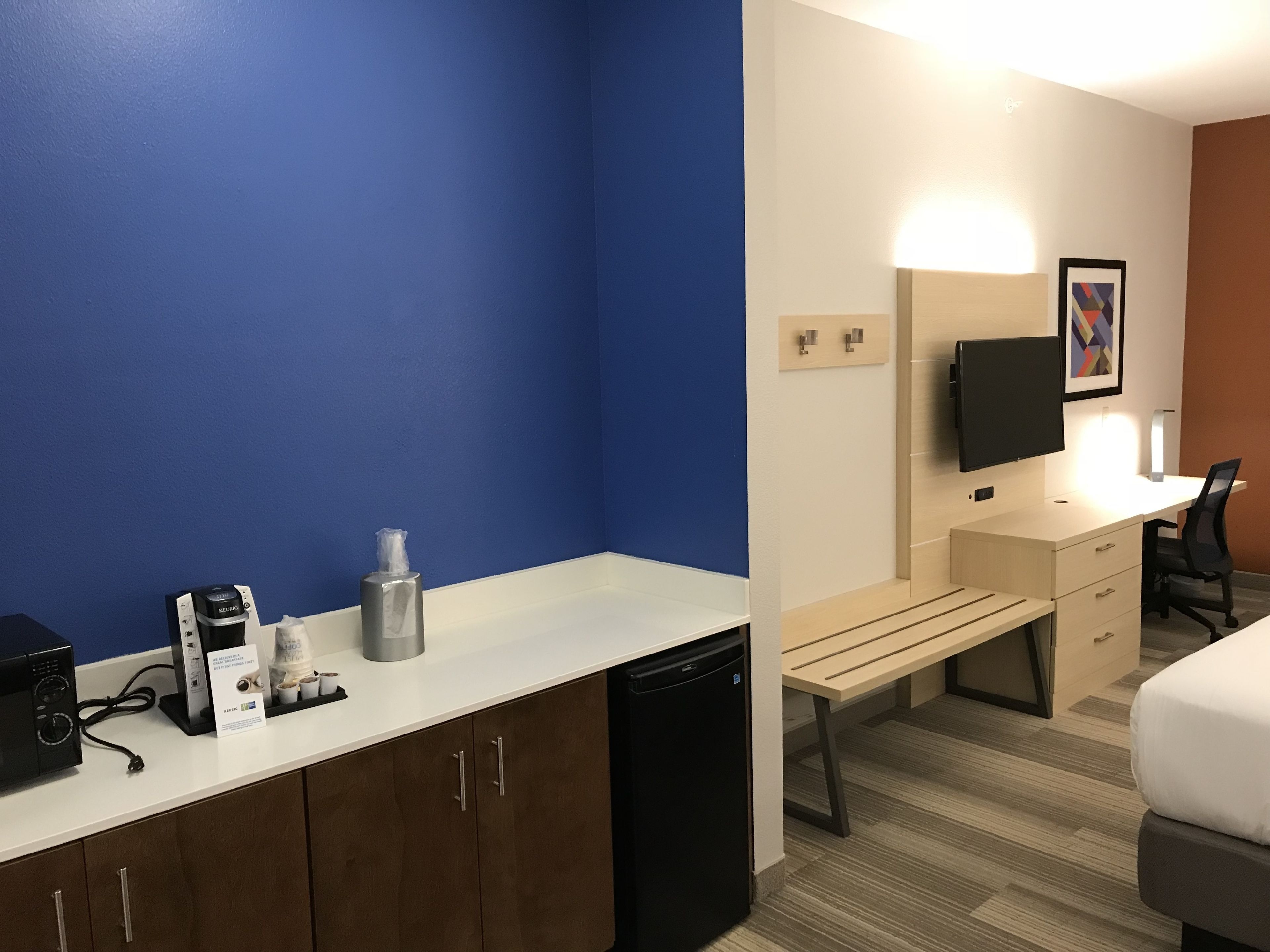 A hotel room with a blue wall, a white desk, a bed, a television, and a coffee machine.