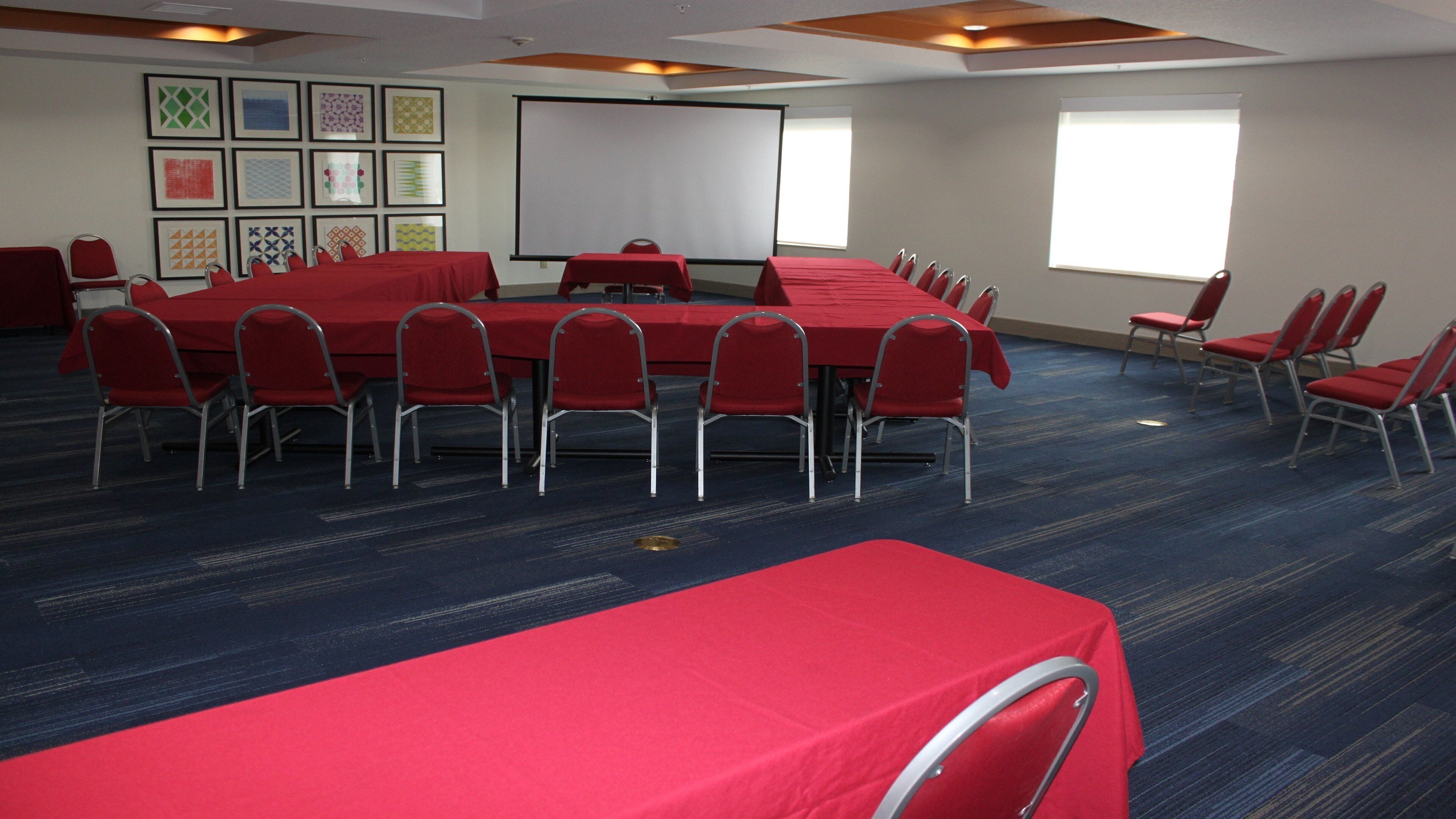A conference room with multiple tables and chairs, a projector screen, and framed artwork on the walls.