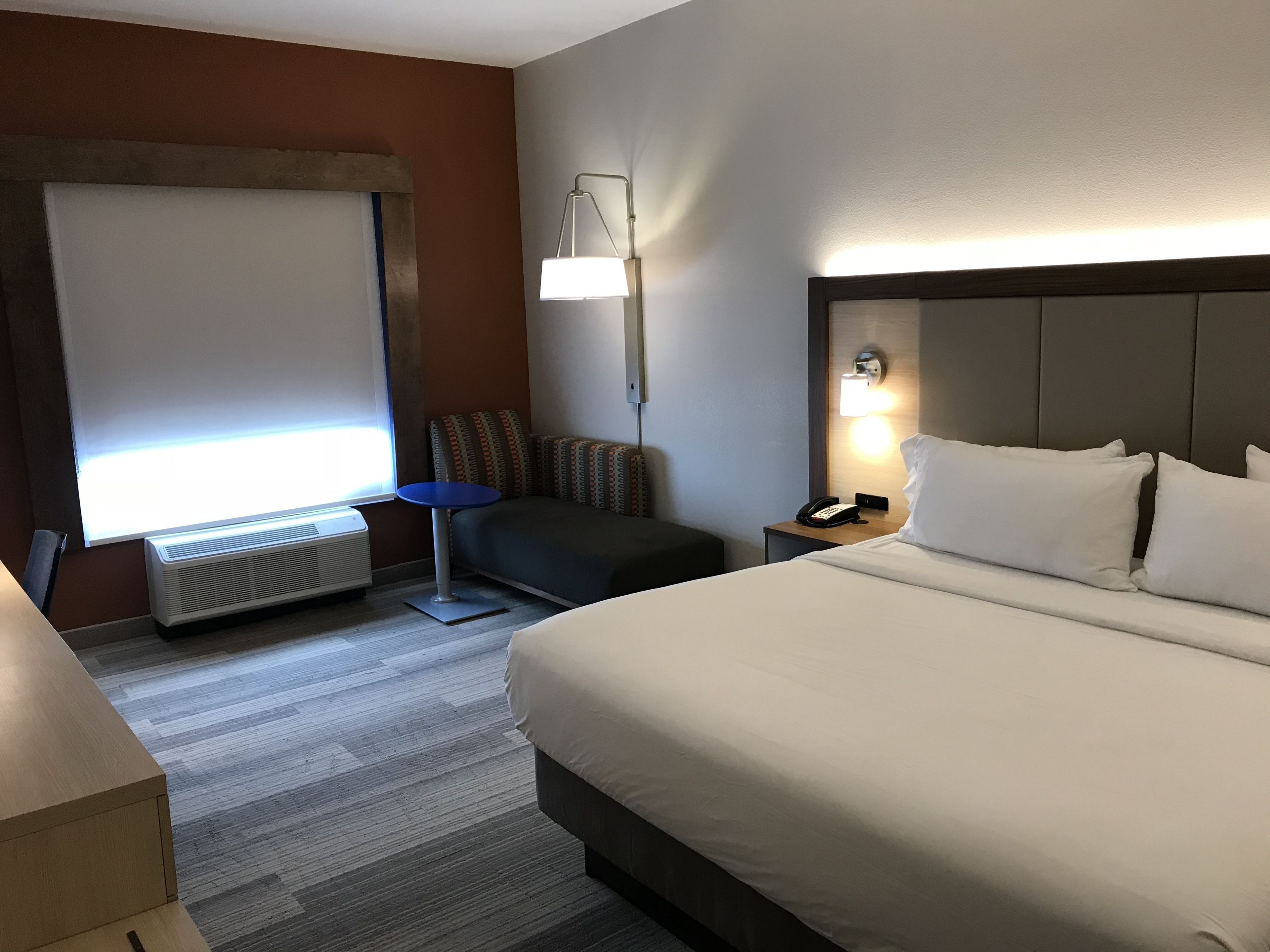 A hotel room with a bed, a desk, a chair, a standing lamp, and a heater.