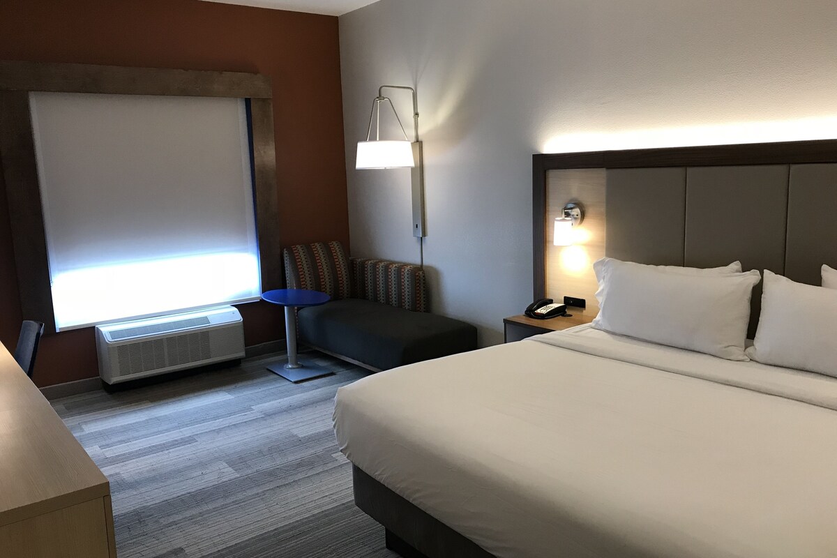 A hotel room with a bed, a desk, a chair, a standing lamp, and a heater.
