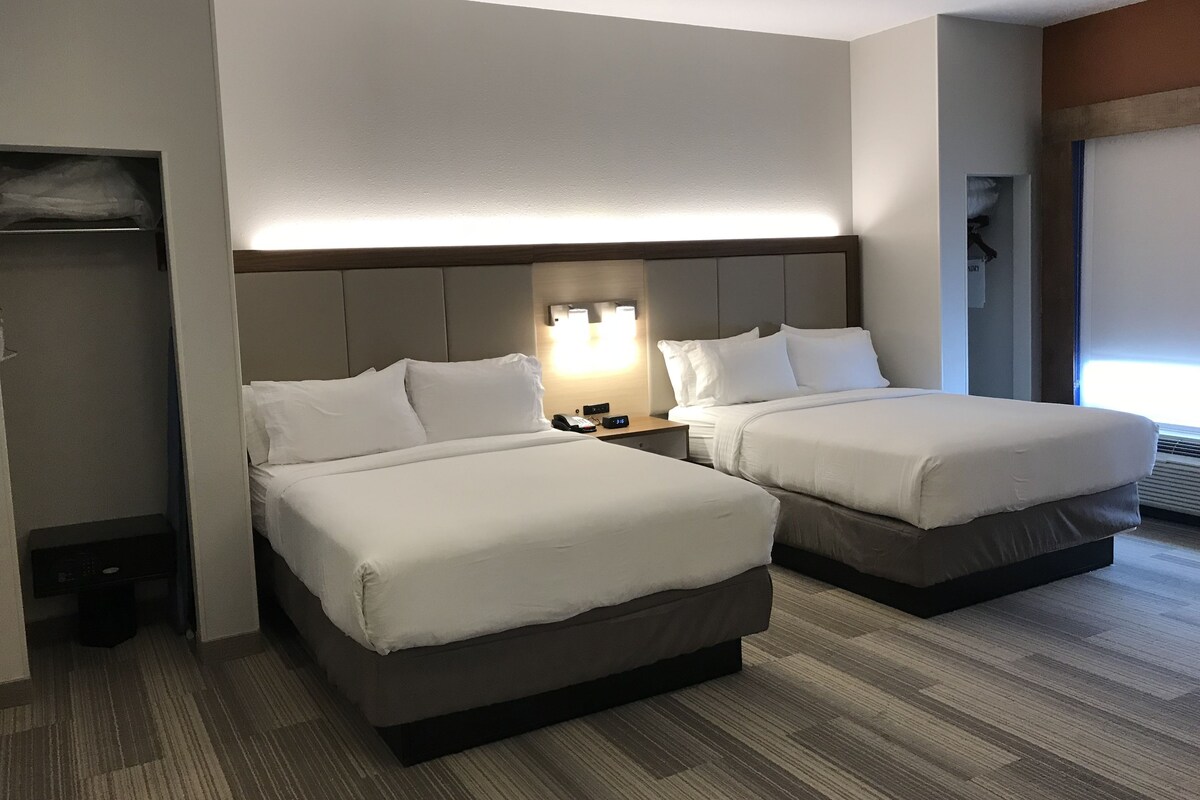 A hotel room with two beds, a nightstand with a lamp, a wardrobe, and a window with blinds.