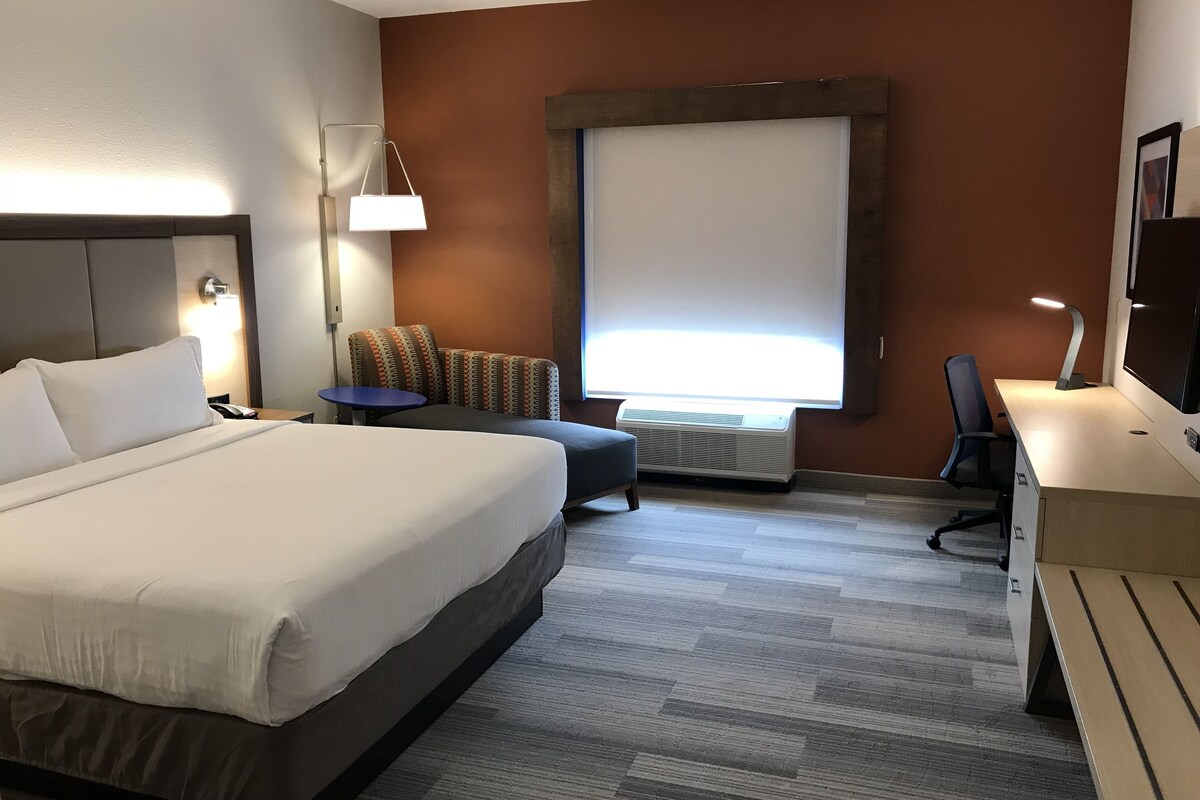 A hotel room with a large bed, a desk, a chair, a small table, and a window with blinds.