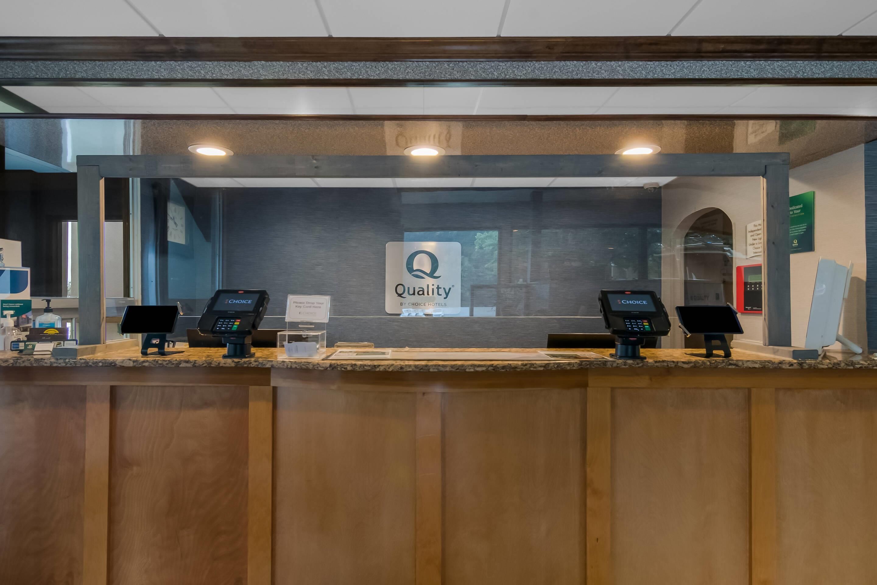 A hotel reception desk with a 'Quality by Choice Hotels' sign, two payment terminals, and a glass partition.