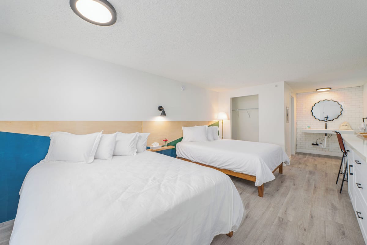 A hotel room with two beds, a desk, a chair, a mirror, and a bathroom with a sink and toilet.