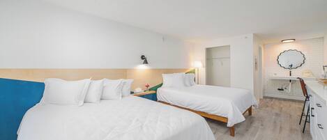 Habitación de hotel con dos camas, un escritorio, una silla, un espejo y un baño con lavamanos e inodoro.