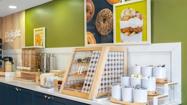A breakfast bar with a coffee machine, a fruit display, and a framed picture of donuts. - Spark by Hilton Murfreesboro (Murfreesboro)