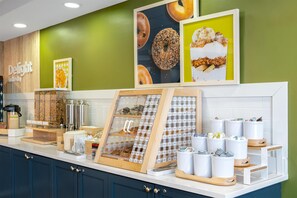 Una barra de desayuno con una máquina de café, una exhibición de frutas y un cuadro con una imagen de rosquillas.