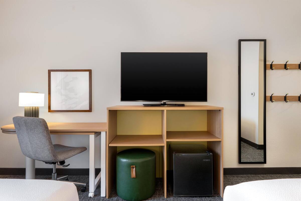 A hotel room with a desk, chair, television, small refrigerator, trash bin, and a mirror on the wall.