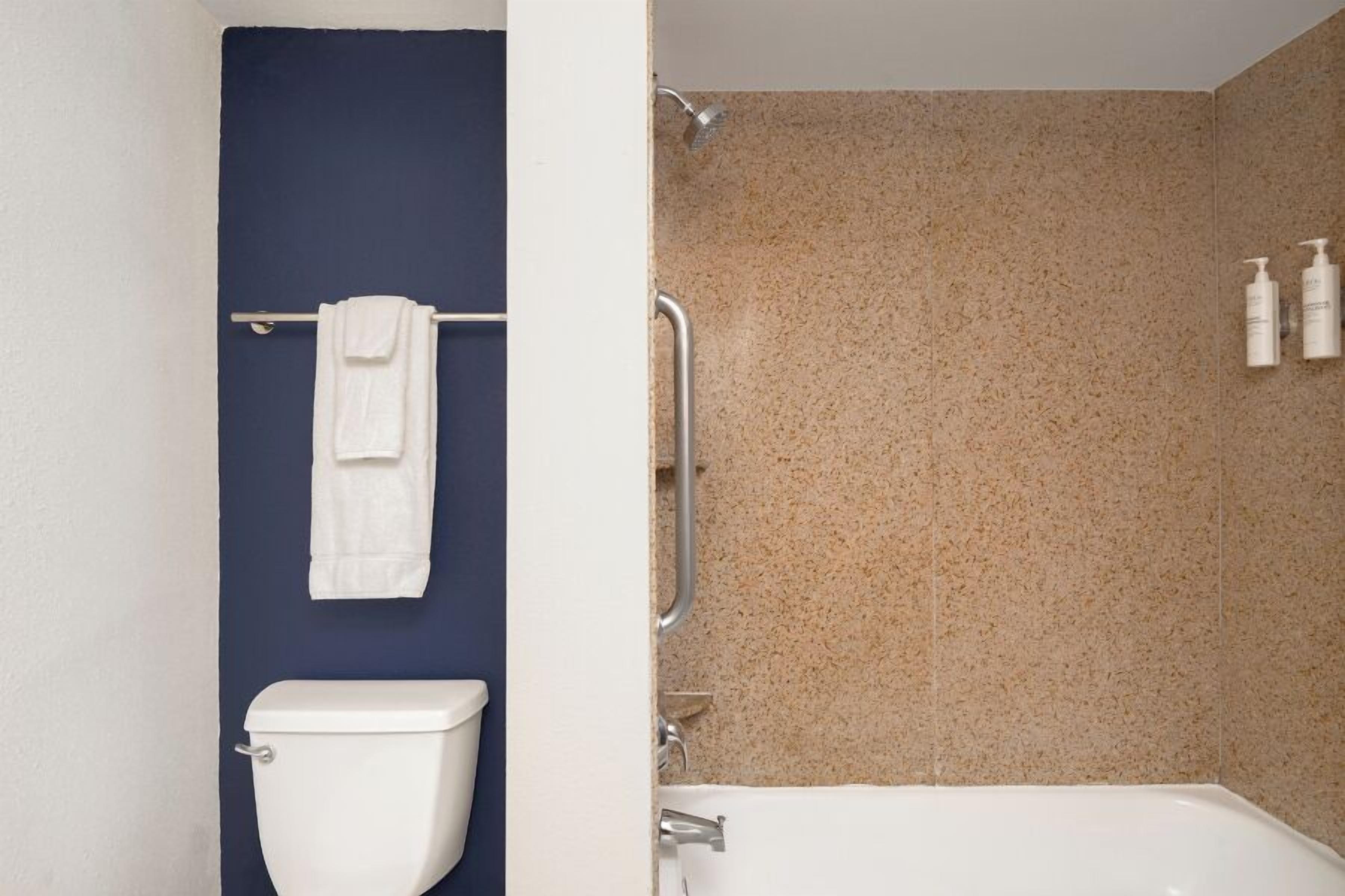 A bathroom with a white toilet, a towel rack with a white towel, a shower area with a beige speckled wall, and two bottles of toiletries on the wall.
