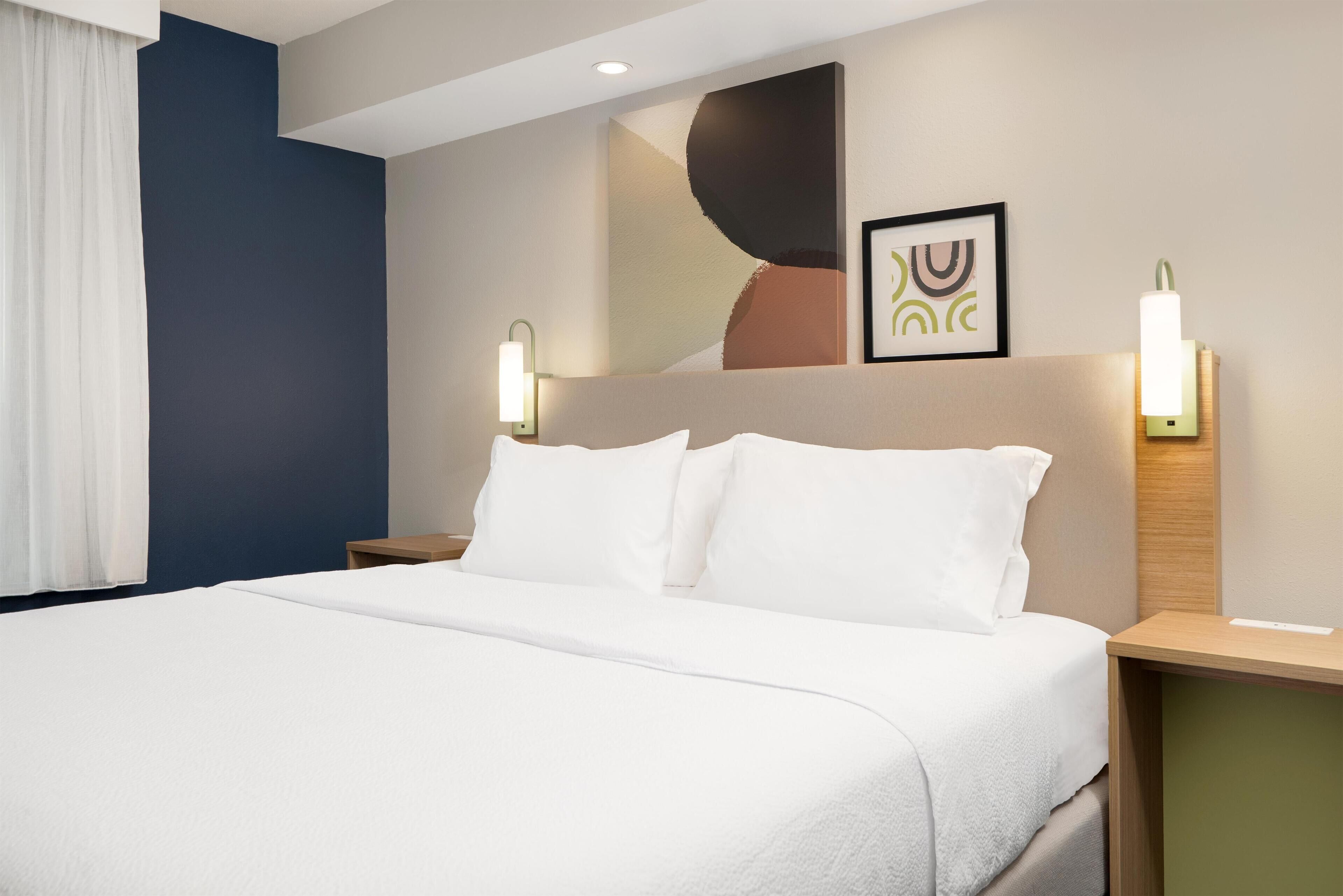 A hotel room with a large bed, two bedside tables, a headboard with artwork, and wall-mounted lights.