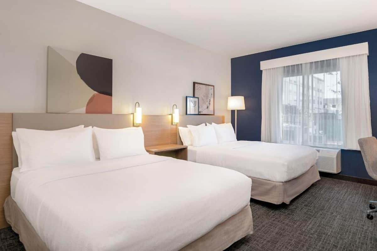 A hotel room with two beds, a desk, a chair, a large window with white curtains, and wall art.