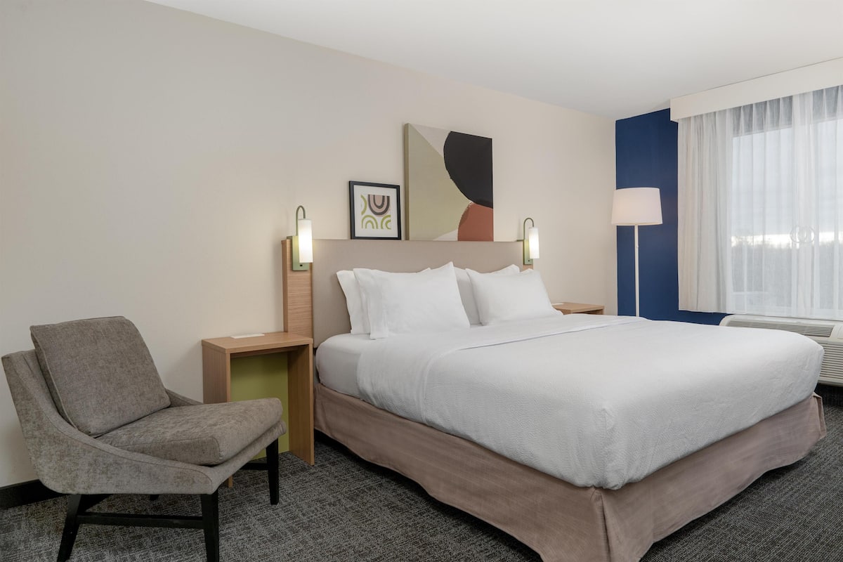 A hotel room with a bed, a chair, a nightstand, and a wall-mounted lamp.
