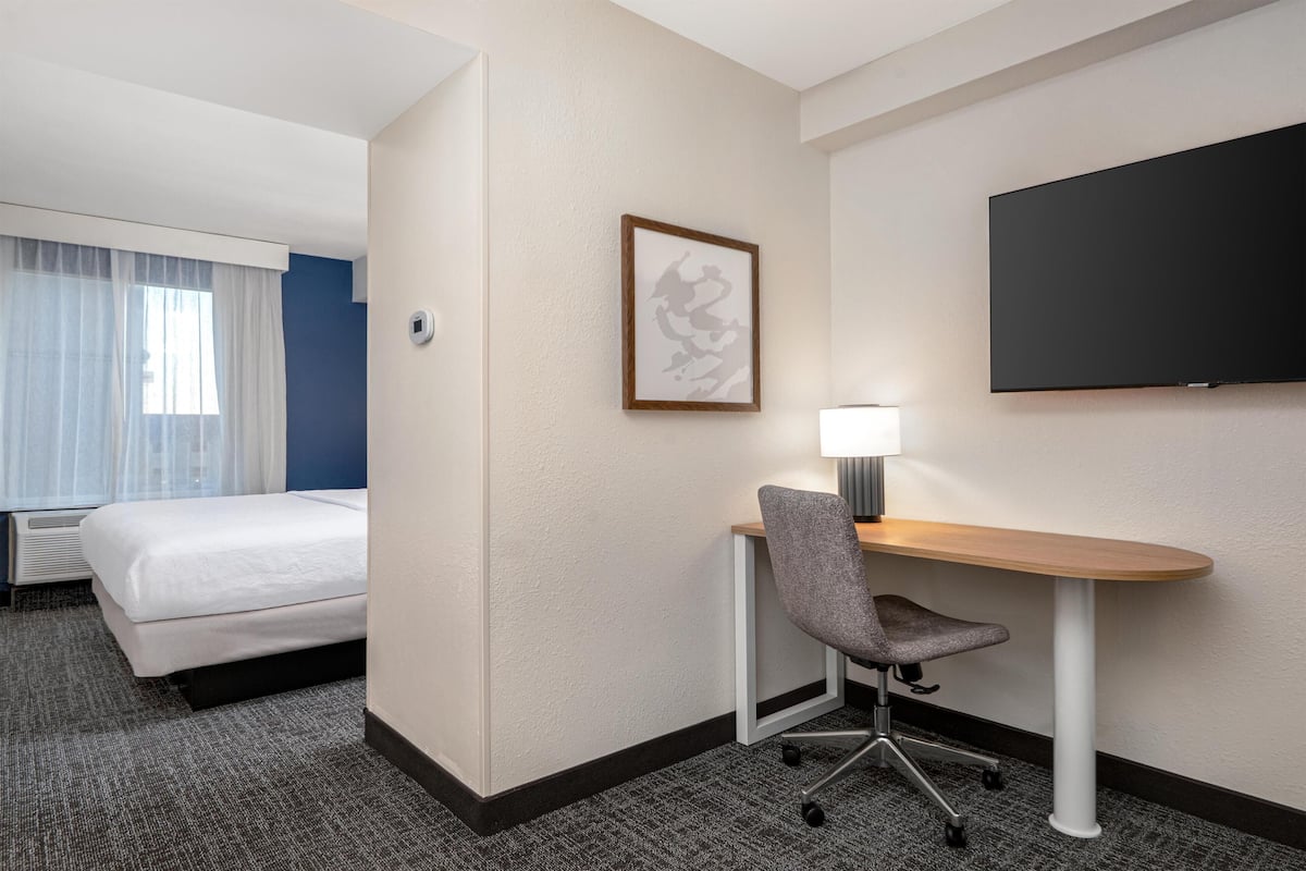A hotel room with a bed, a desk with a chair, a flat-screen TV, and a framed picture on the wall.