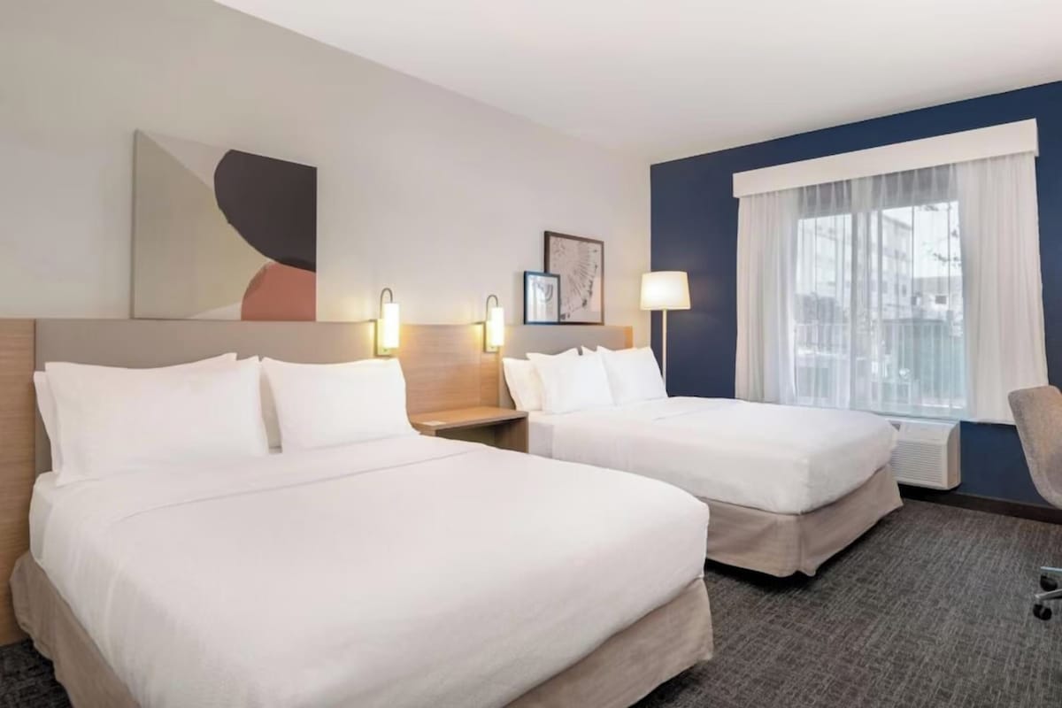 A hotel room with two beds, a nightstand, a lamp, a chair, and a large window with curtains.