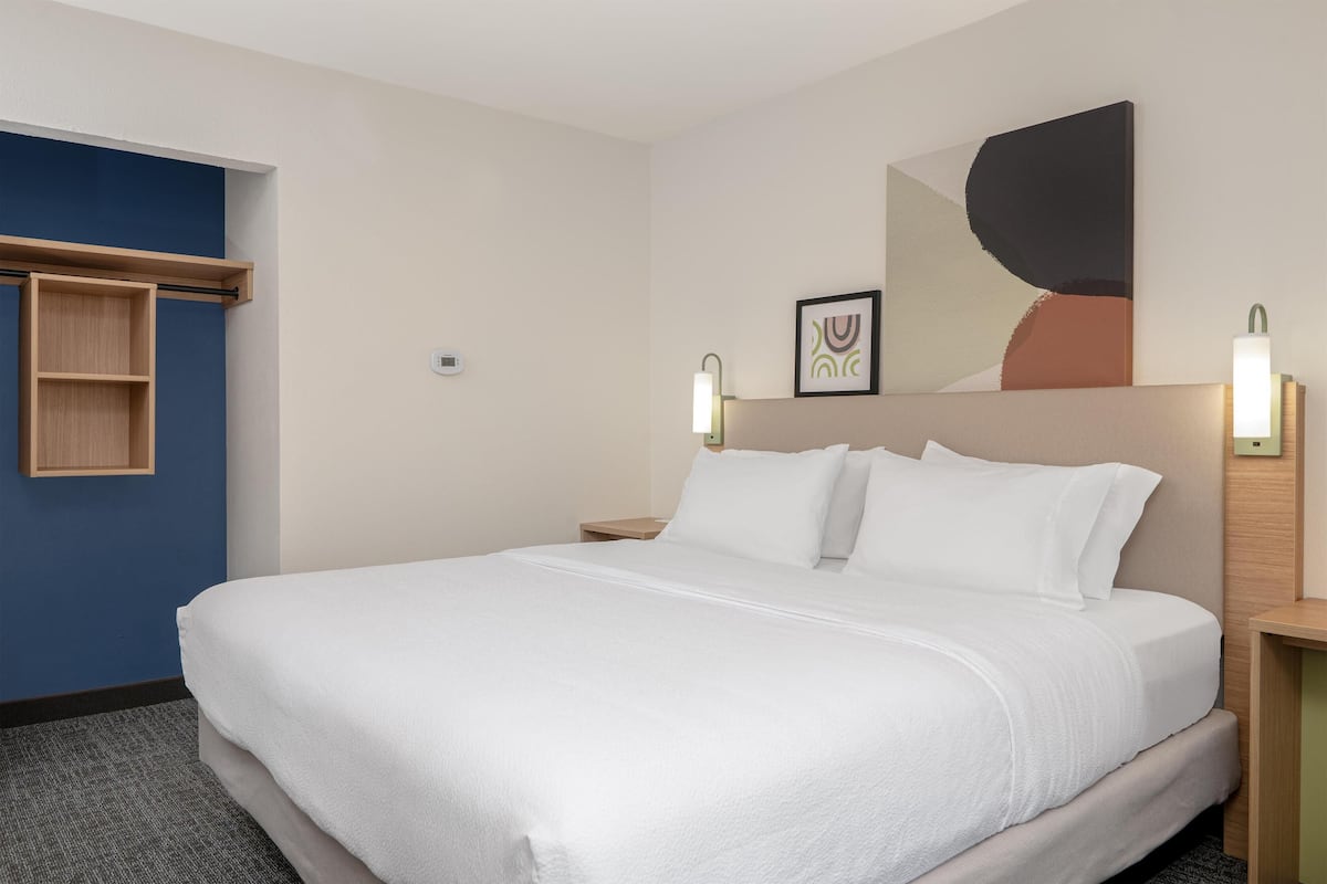 A hotel room with a large bed, a wooden shelf, a wall-mounted artwork, and two bedside lamps.