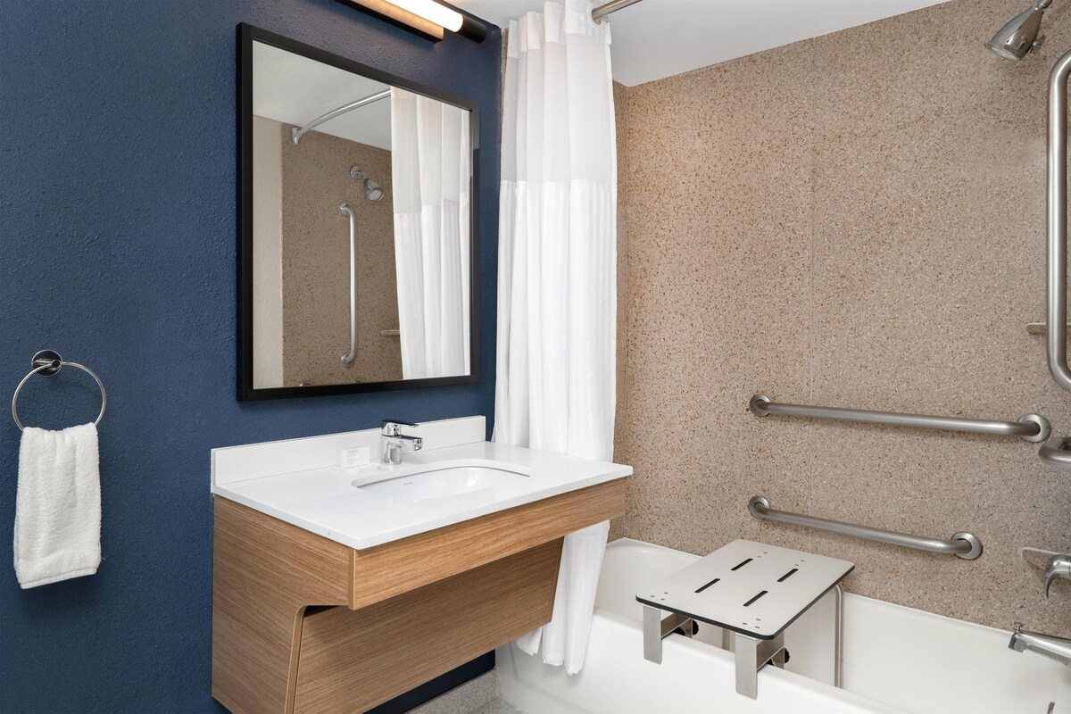 Ein modernes Badezimmer mit einem weißen Waschbecken, einem Holz-Badmöbel, einem Spiegel, einer Dusche mit Haltegriffen und einem Handtuchhalter.